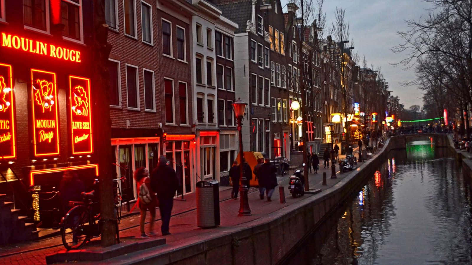 Red Light District 桌布 壁紙 — 阿姆斯特丹的暮色運河，發光的紅色霓虹燈招牌倒映在水面上，行人沿著鵝卵石小徑漫步。