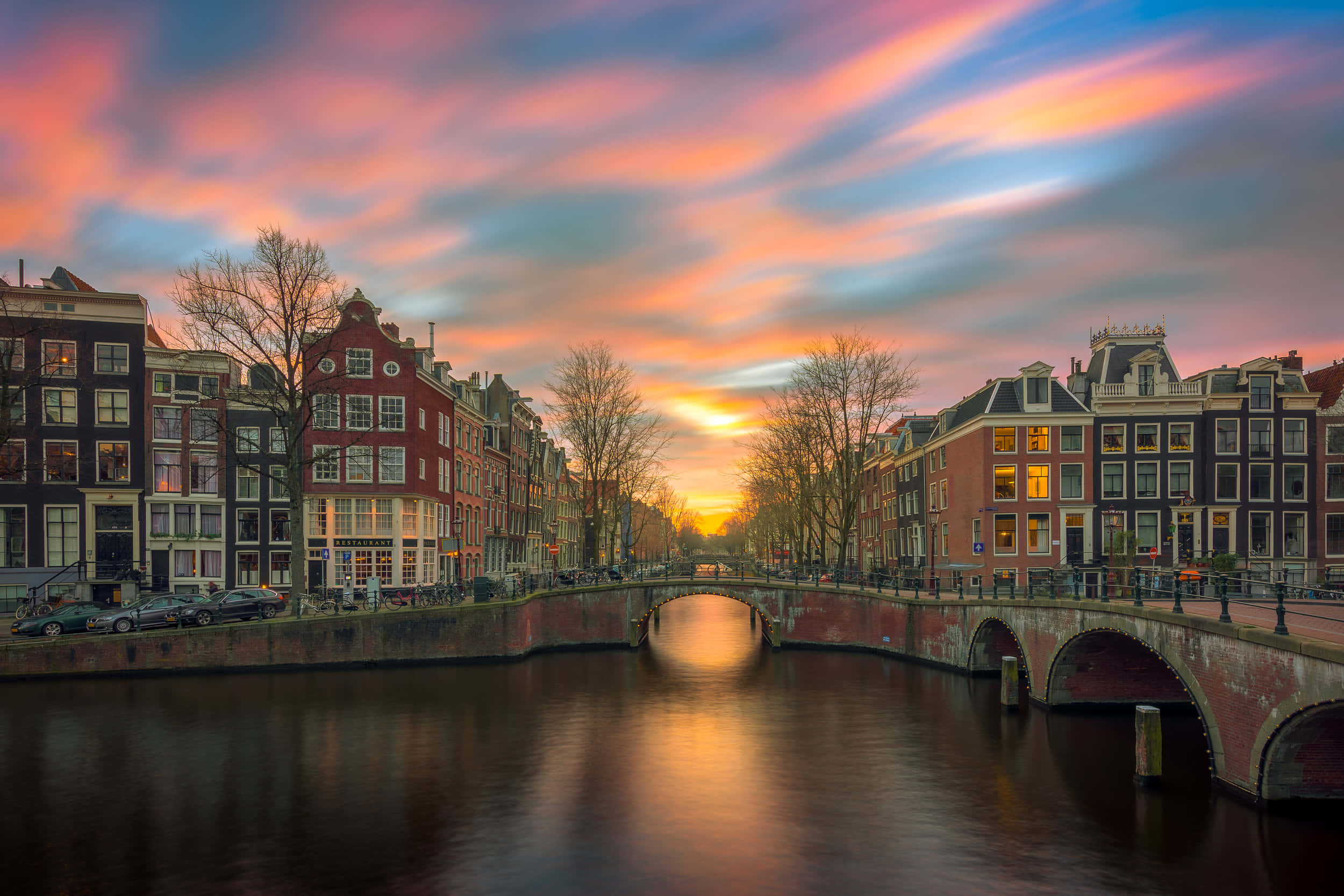 Netherlands 桌布 壁紙 — 阿姆斯特丹運河上壯麗的日落，色彩繽紛的雲彩倒映在平靜的水面和歷史悠久的橋樑上。