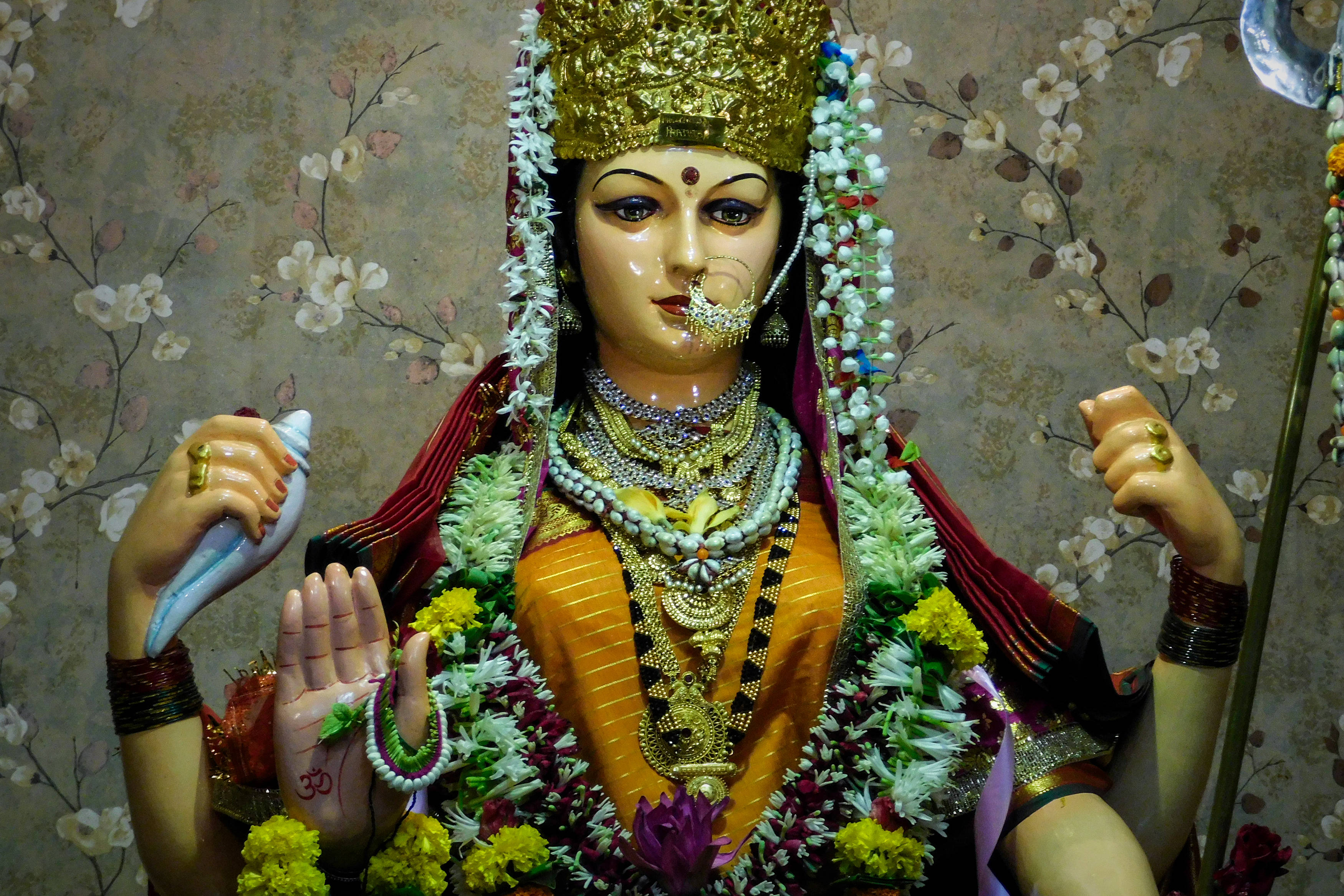 阿姆比瑪女神納瓦拉提 壁紙 — 一尊裝飾華麗的印度教女神雕像，戴著金色皇冠珠寶、花環和鮮豔的橙色服飾，背景是花卉牆紙。