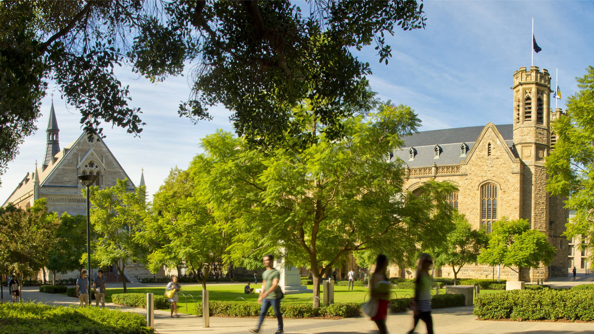 阿德萊德大學建築風景 壁紙 — 學生們在陽光明媚的校園廣場漫步，四周是茂密的綠樹和歷史悠久的石砌建築，頭頂是清澈的藍天。