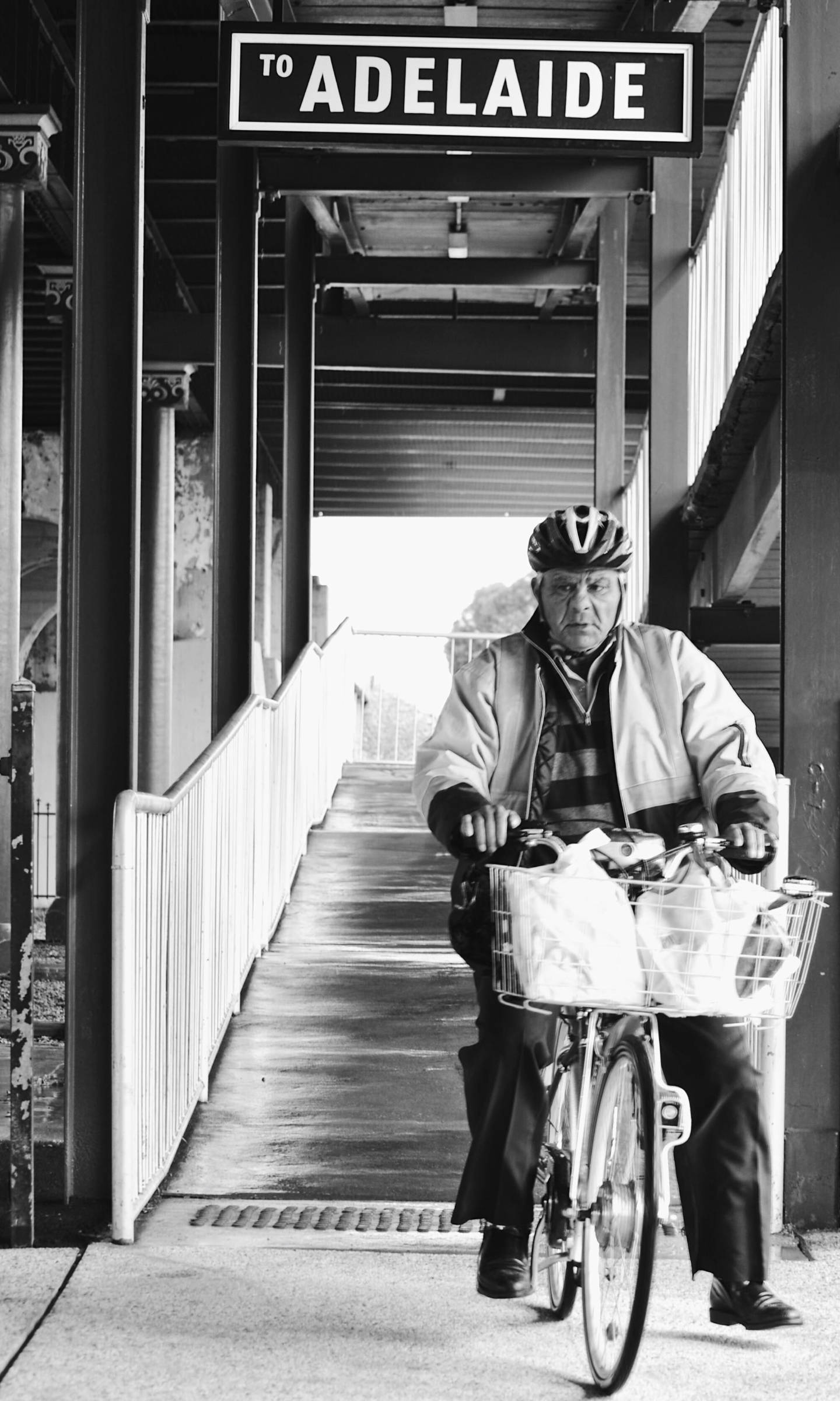 阿德萊德路牌下騎自行車的長者 壁紙 — 一位戴頭盔的老年單車手，在寫有「TO ADELAIDE」黑白字樣的有蓋車站月台下，騎著帶籃子的單車。
