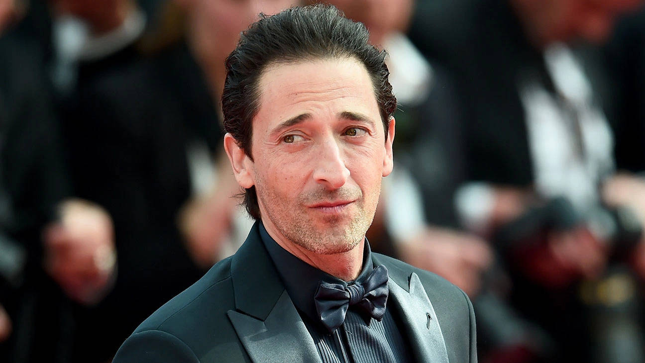 阿德里安·布洛迪全黑正裝照 壁紙 — Adrien Brody 身穿黑色燕尾服配蝴蝶結，在紅地毯上若有所思地凝視鏡頭，身後是模糊的人群。