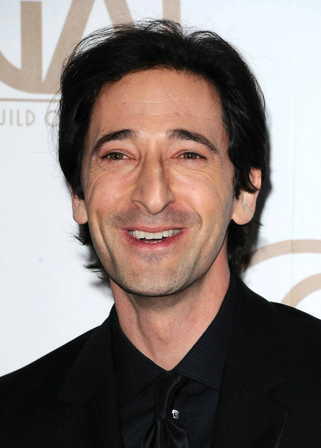 阿德里安·布洛迪微笑肖像照 壁紙 — Adrien Brody 身穿黑色西裝，在金色字體的白色活動背景前溫暖地微笑。
