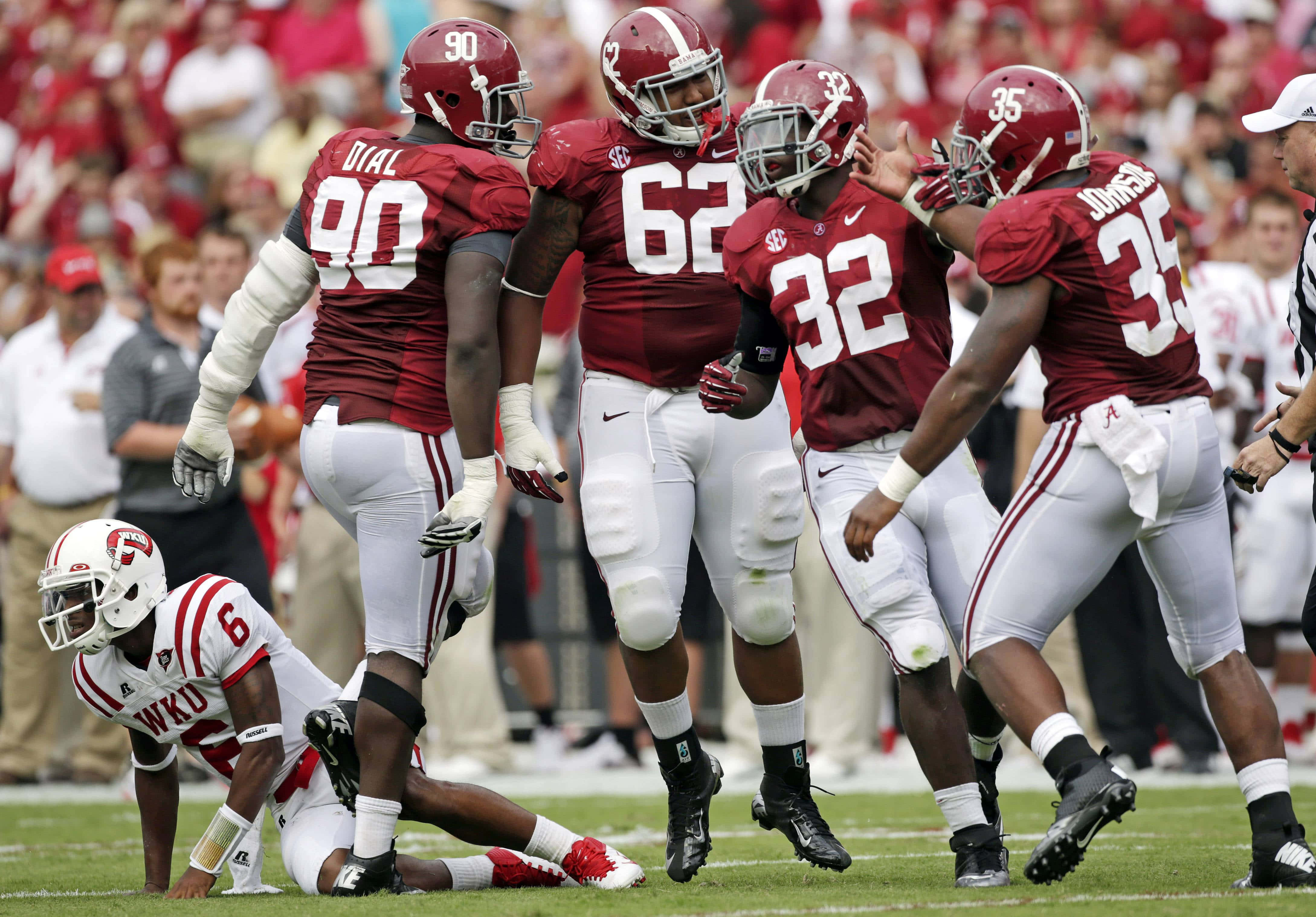Roll Tide! 壁紙 — 阿拉巴馬大學美式足球員身穿深紅色和白色裝備，在陽光明媚的球場上慶祝一次進攻，附近有一名倒下的WKU球員。