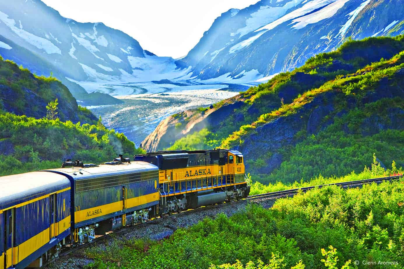 阿拉斯加安克雷奇火車與綠山風景 壁紙 — 一輛鮮黃色的阿拉斯加鐵路火車蜿蜒穿過翠綠的山谷，兩旁是白雪覆蓋的山脈和遠處的冰川。