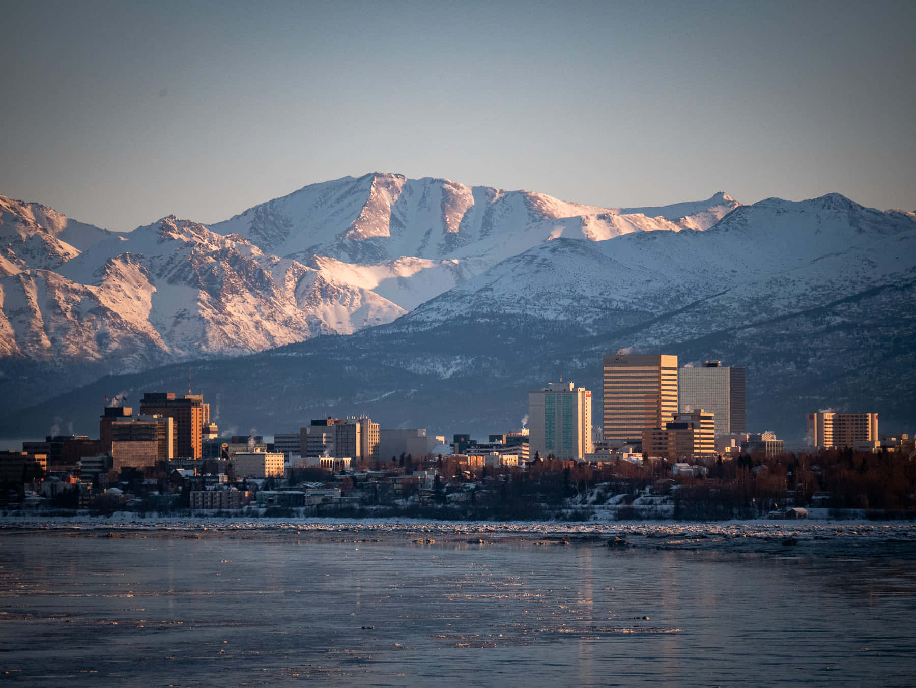 阿拉斯加安克雷奇雪峰城市風景 壁紙 — 積雪的山峰在金色日出光芒中閃耀，背後是城市天際線倒映在部分結冰的河流中。