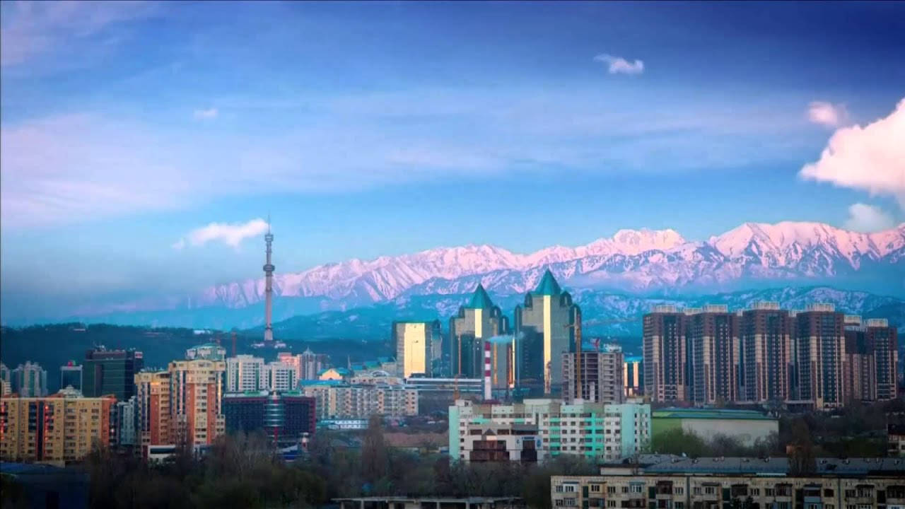 阿拉木圖全景 壁紙 — 城市景觀，現代建築與白雪覆蓋的山脈在藍天白雲下。