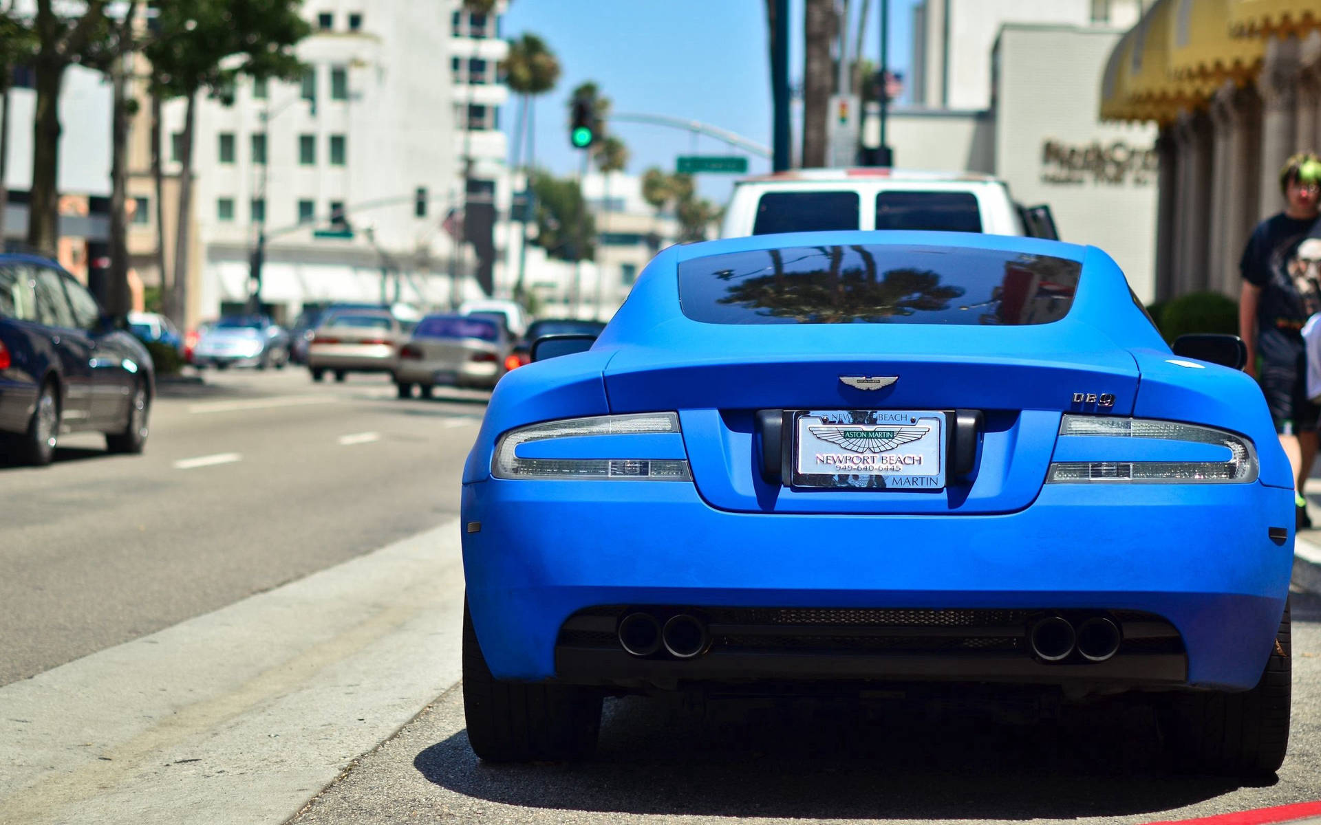 藍色Aston Martin DB9停泊在帶有棕櫚樹和建築物的城市街道上。