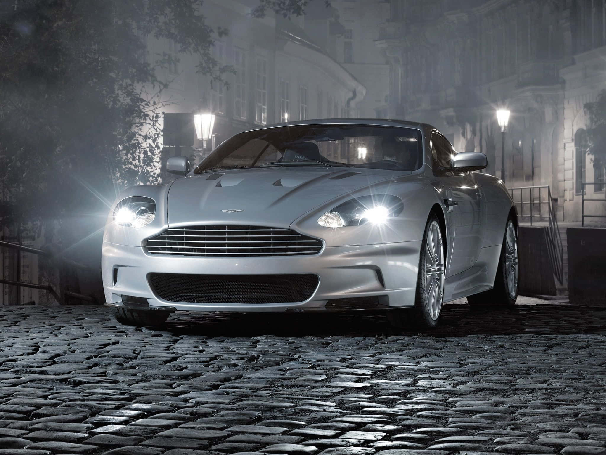 阿斯頓·馬丁DB9——經典永恆 壁紙 — 一輛銀色Aston Martin跑車在夜間霧濛濛的鵝卵石街道上，在街燈下閃閃發光。
