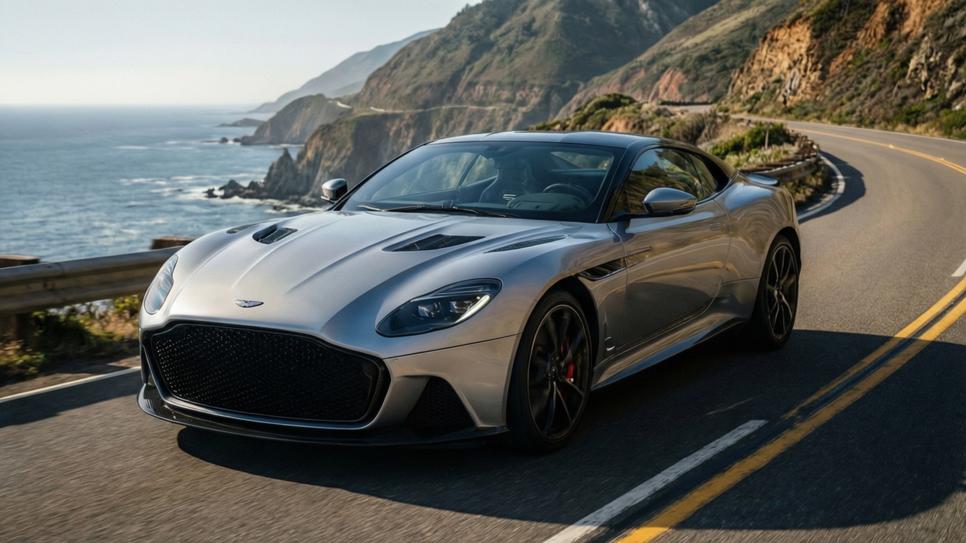 銀色 Aston Martin 跑車在海洋懸崖附近的蜿蜒海岸道路上行駛。