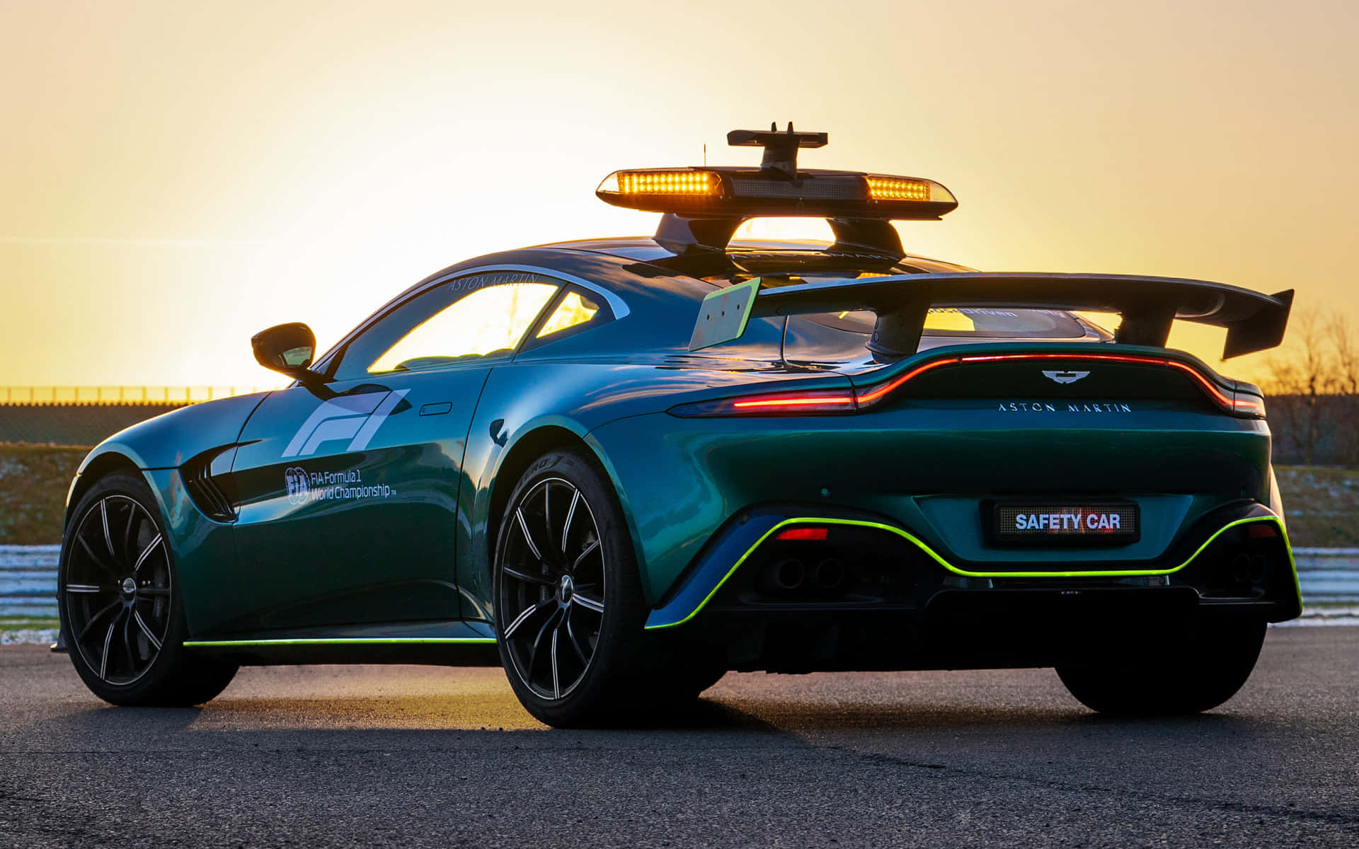 阿斯頓·馬丁的性能藝術 壁紙 — 一架時尚的綠色 Aston Martin 安全車，閃爍著燈光，停泊在金色日落下的賽道上。