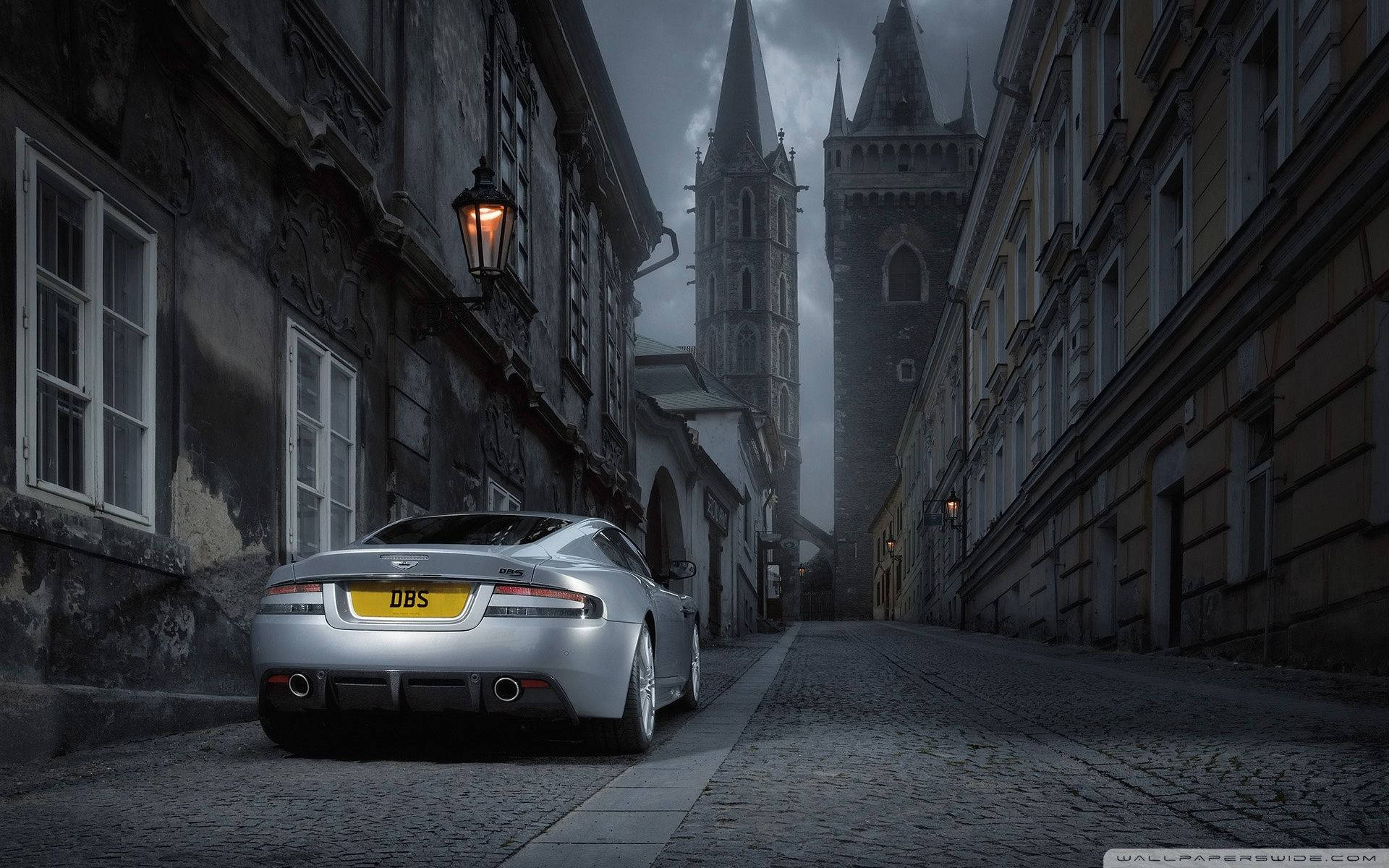銀色Aston Martin DBS夜間停泊在歷史建築附近的狹窄鵝卵石街道上。