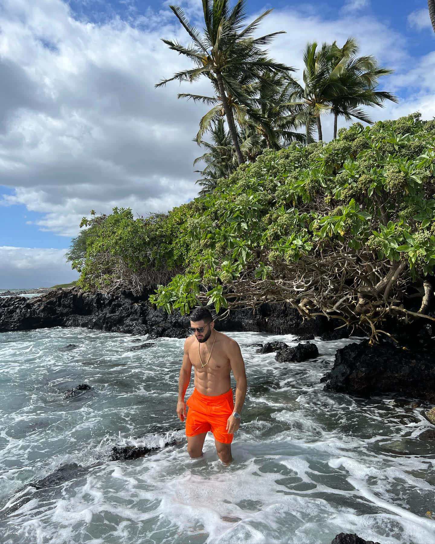 穿著橙色泳褲的男子在熔岩岩石和棕櫚樹附近的海浪中涉水。