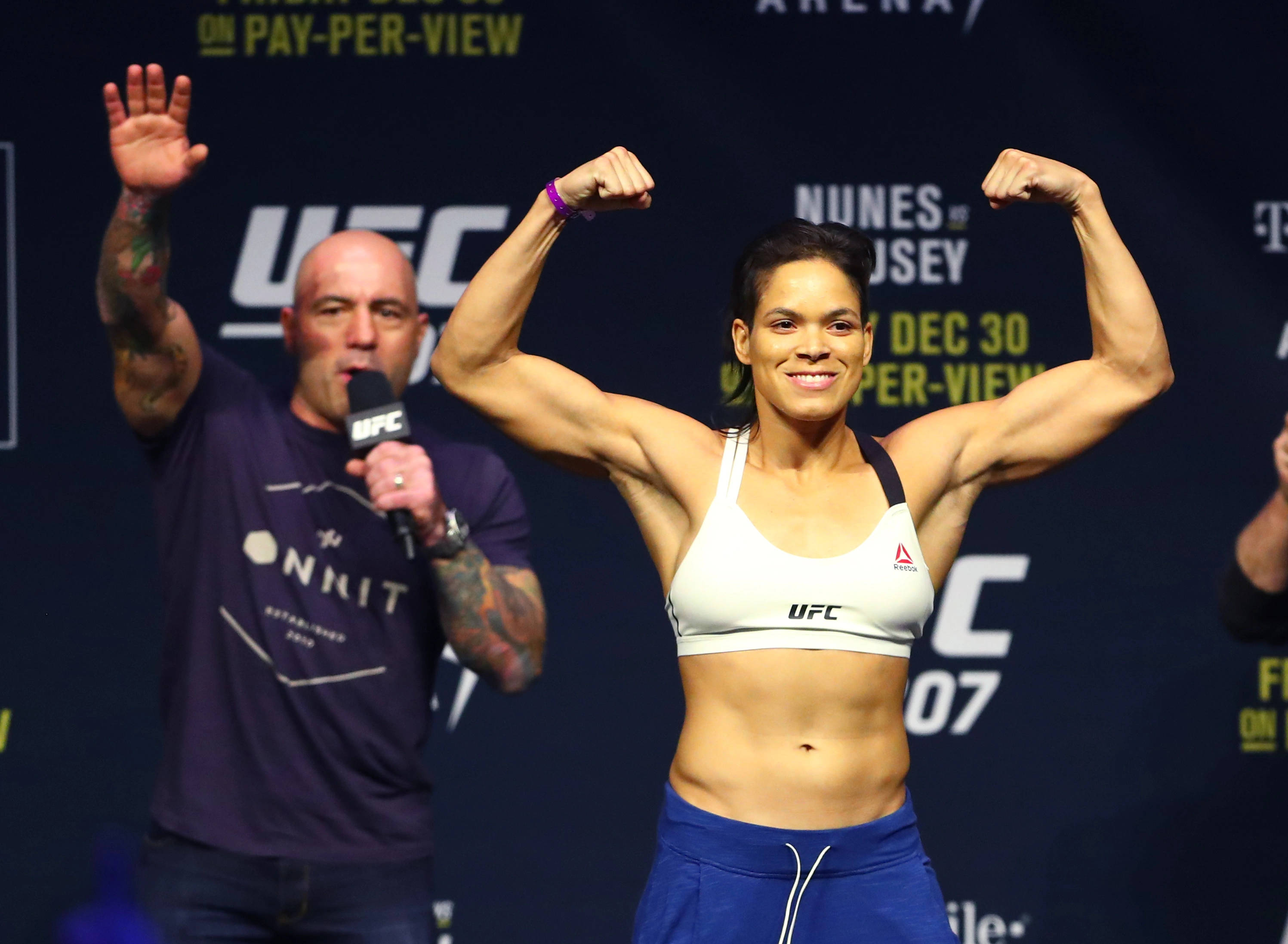 阿曼達·努內斯——無敵UFC冠軍 壁紙 — 一位女性UFC拳手在稱重活動中自信地微笑，彎曲她肌肉發達的手臂，而一位有紋身的男子在她旁邊拿著麥克風。