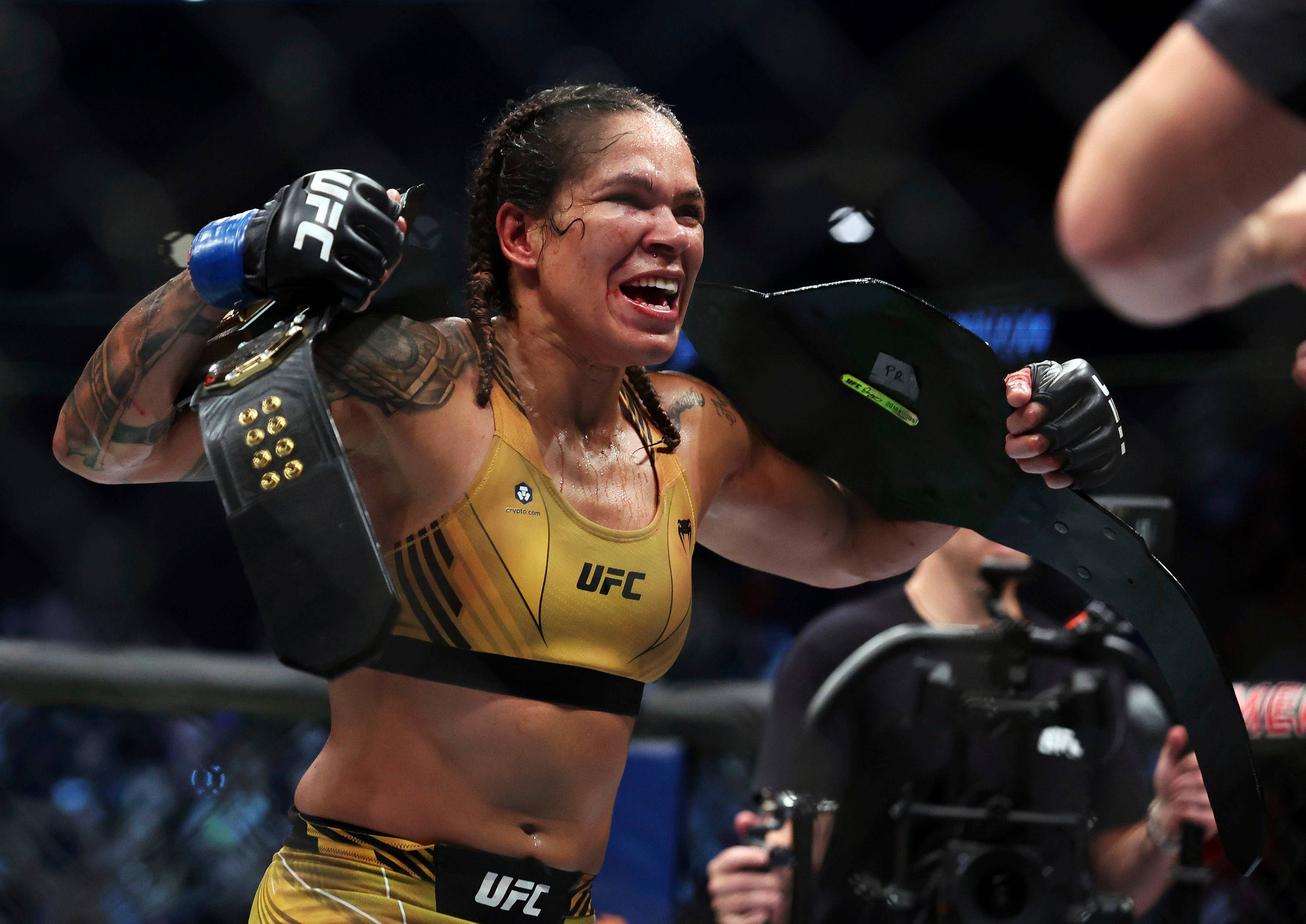 阿曼達·努內斯勝利冠軍 壁紙 — 一位身穿金色制服、勝利的女性UFC拳手在八角籠內高舉冠軍腰帶慶祝勝利。