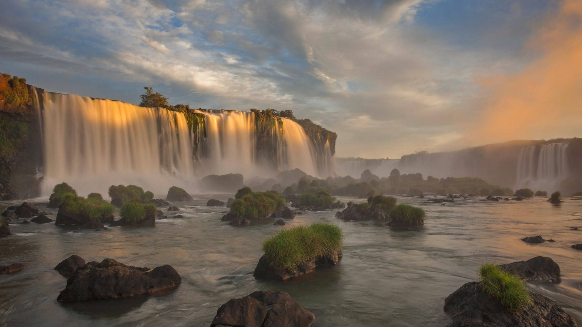 阿根廷伊瓜蘇瀑布 壁紙 — 夕陽以金色光芒沐浴著巨大的伊瓜蘇瀑布，霧氣從流經長滿苔蘚岩石的瀑布水中升起。