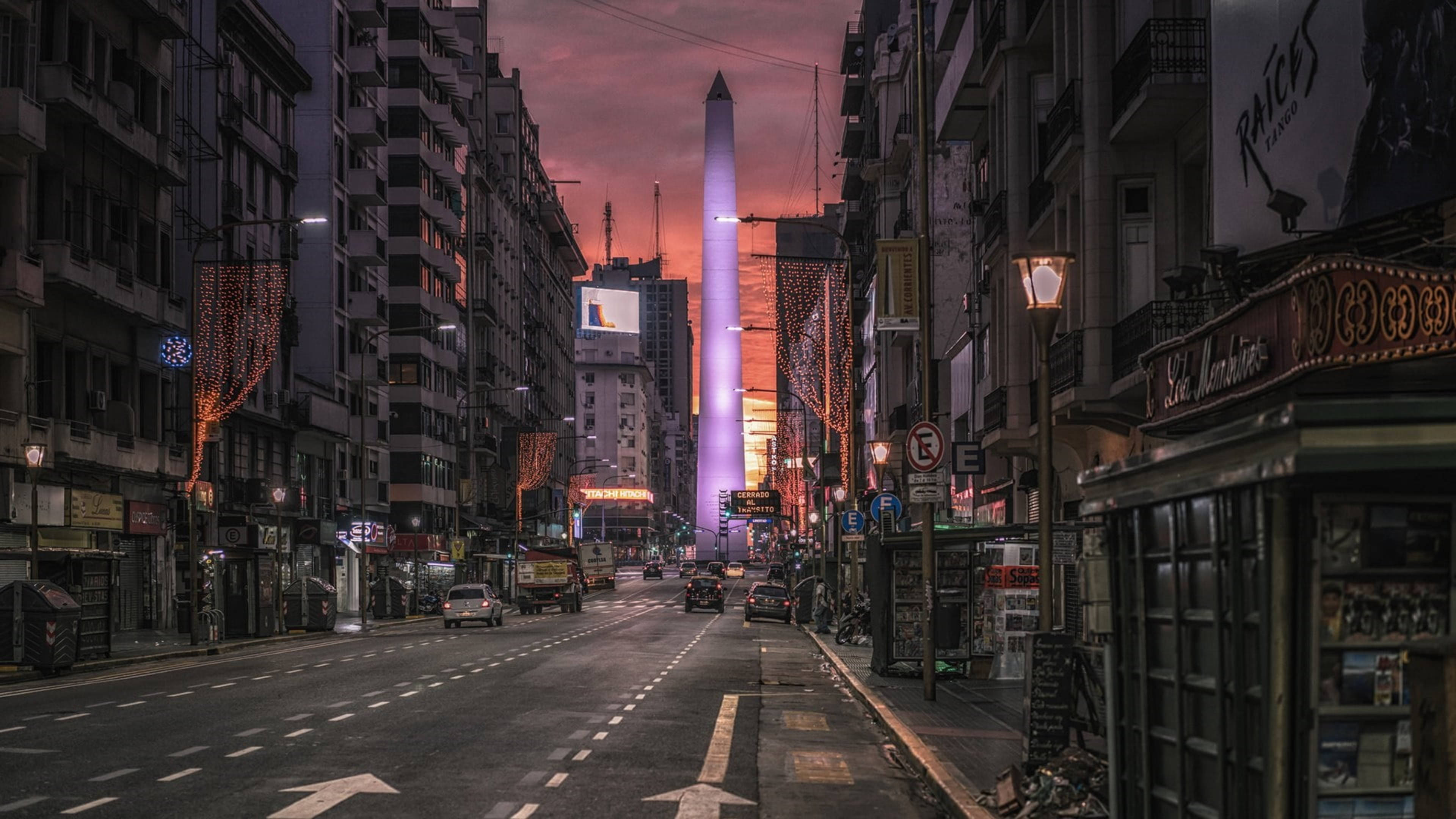 阿根廷方尖碑街景城市風光 壁紙 — 黃昏時分的城市街道，一座發光的方尖碑高聳於建築物之上，建築物之間掛滿節日燈飾。