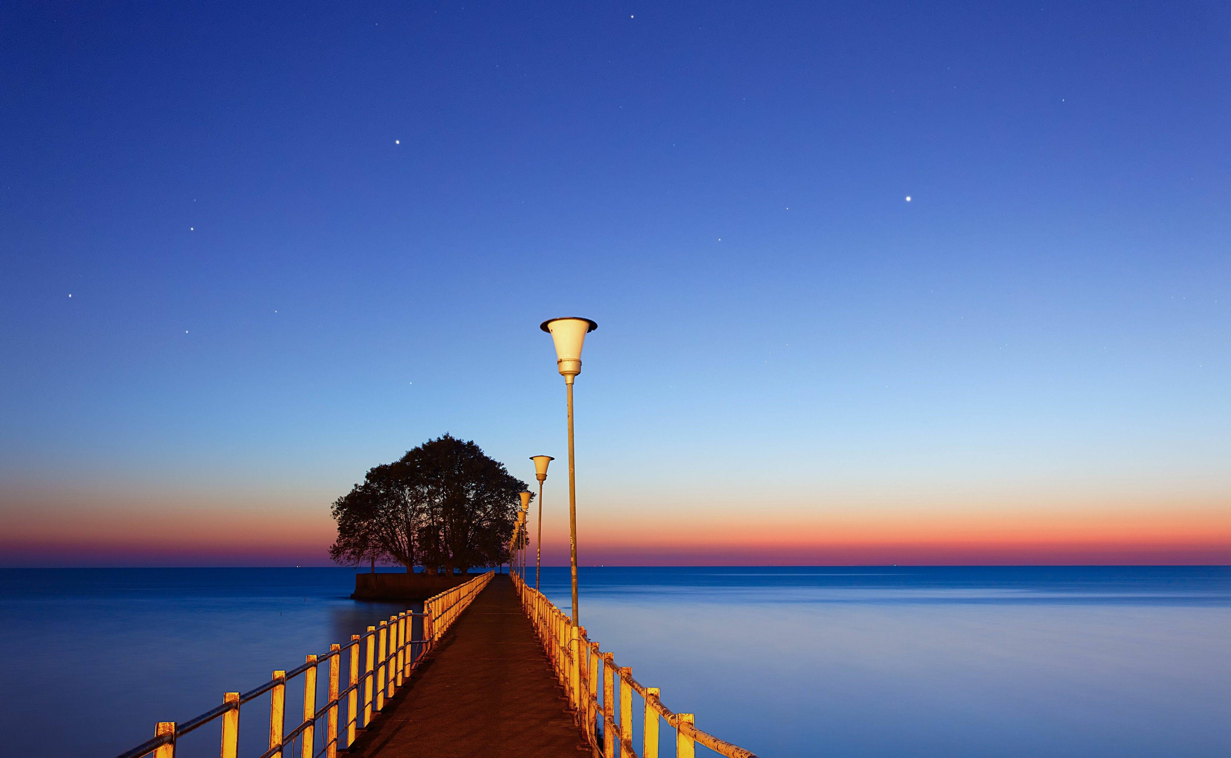 阿根廷海灘夜空 壁紙 — 一條長長的木碼頭延伸至平靜的水面，上方是帶有橙色和深藍色星星的暮色天空。