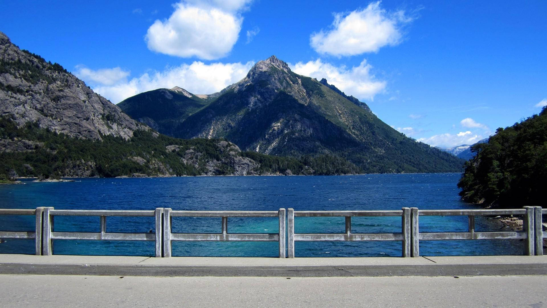 以木製欄杆為框架的綠松石色湖泊景觀，周圍是藍天下的綠色山脈。