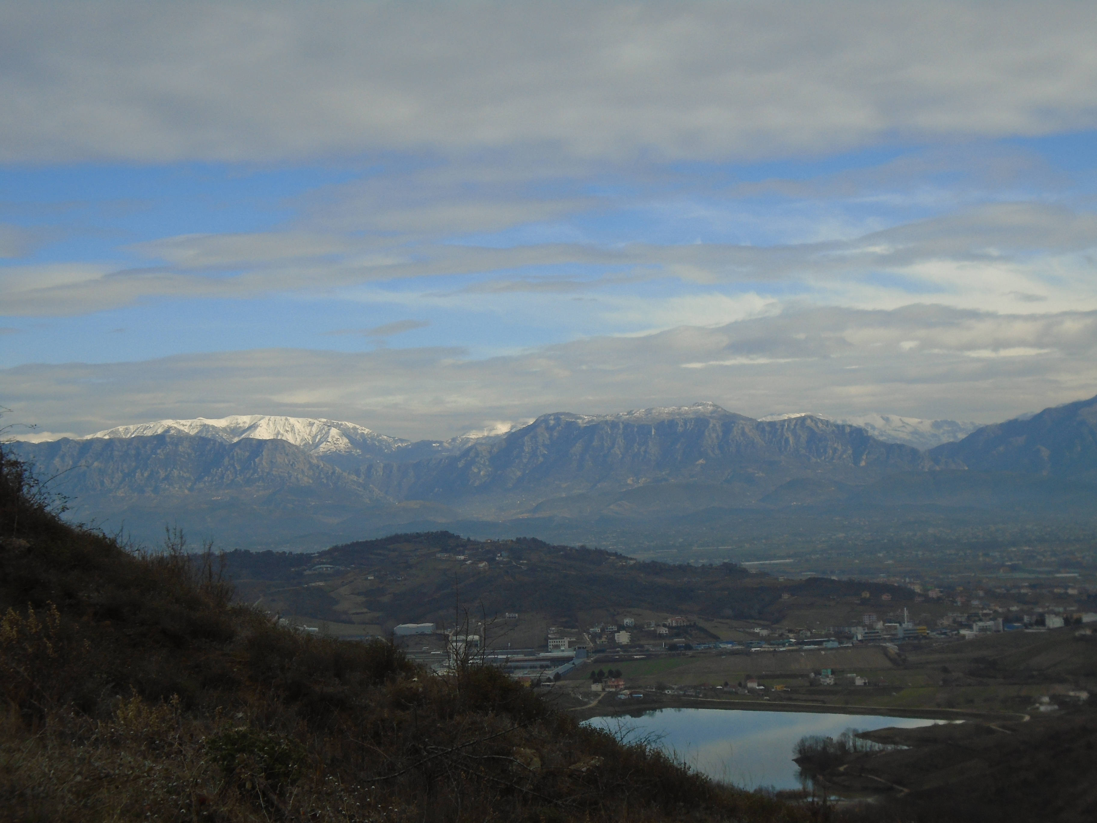 阿爾巴尼亞東部山脈 壁紙 — 白雪覆蓋的山峰在谷城後方升起，平靜的湖泊倒映著多雲的天空。