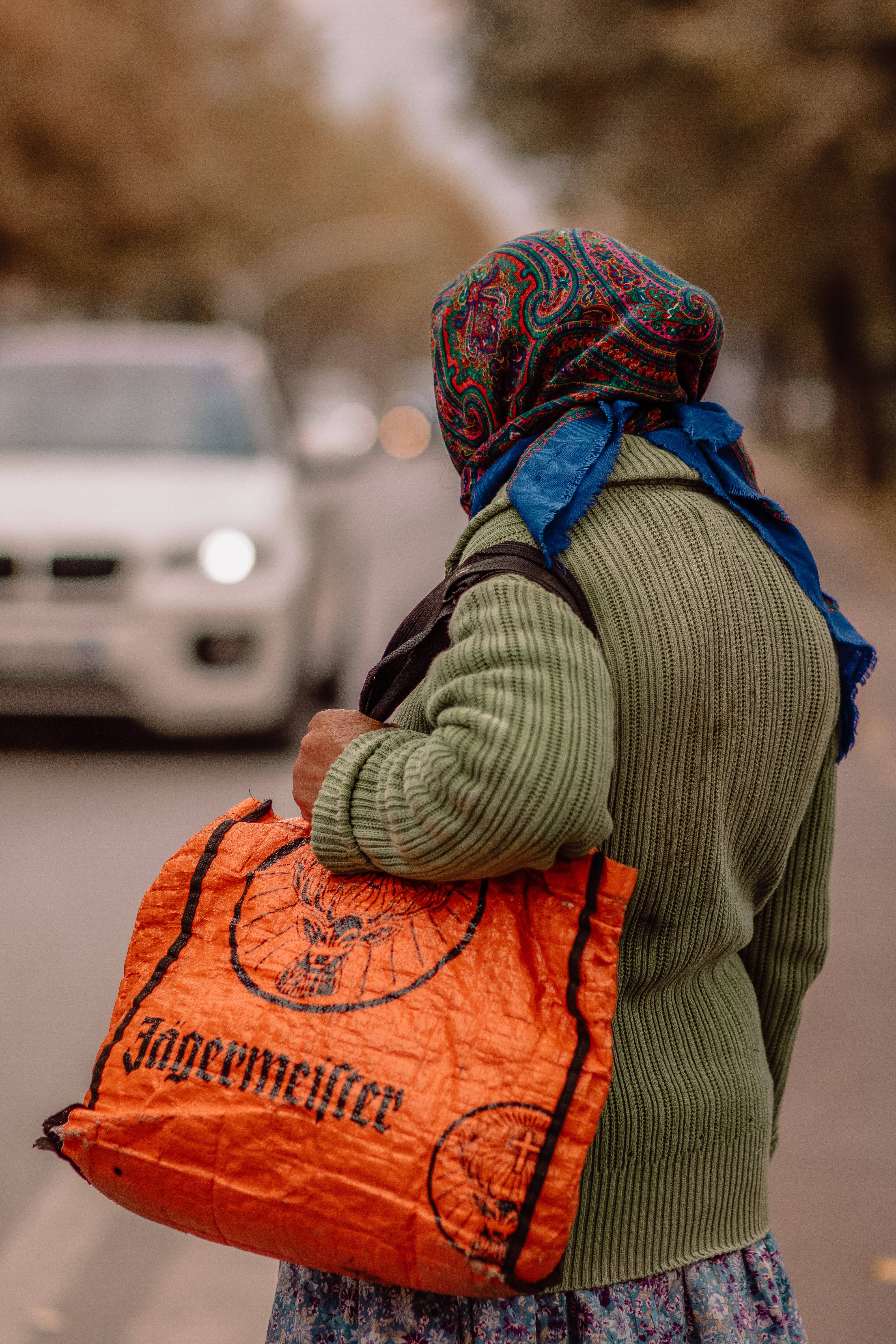 阿爾巴尼亞老婦人 壁紙 — 一個人戴著彩色頭巾、穿著綠色毛衣，背著橙色Jägermeister袋，沿著街道行走，背景有一輛白色汽車。