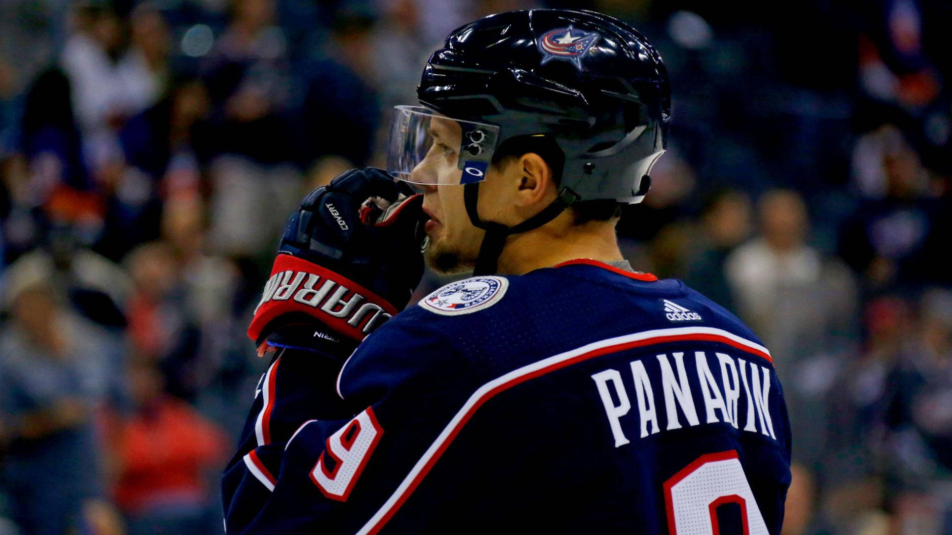 阿特米·帕納林嗅聞手套 壁紙 — 冰球運動員 Panarin 身穿深藍色球衣（9號），戴著頭盔和手套，在冰場上微微轉頭。