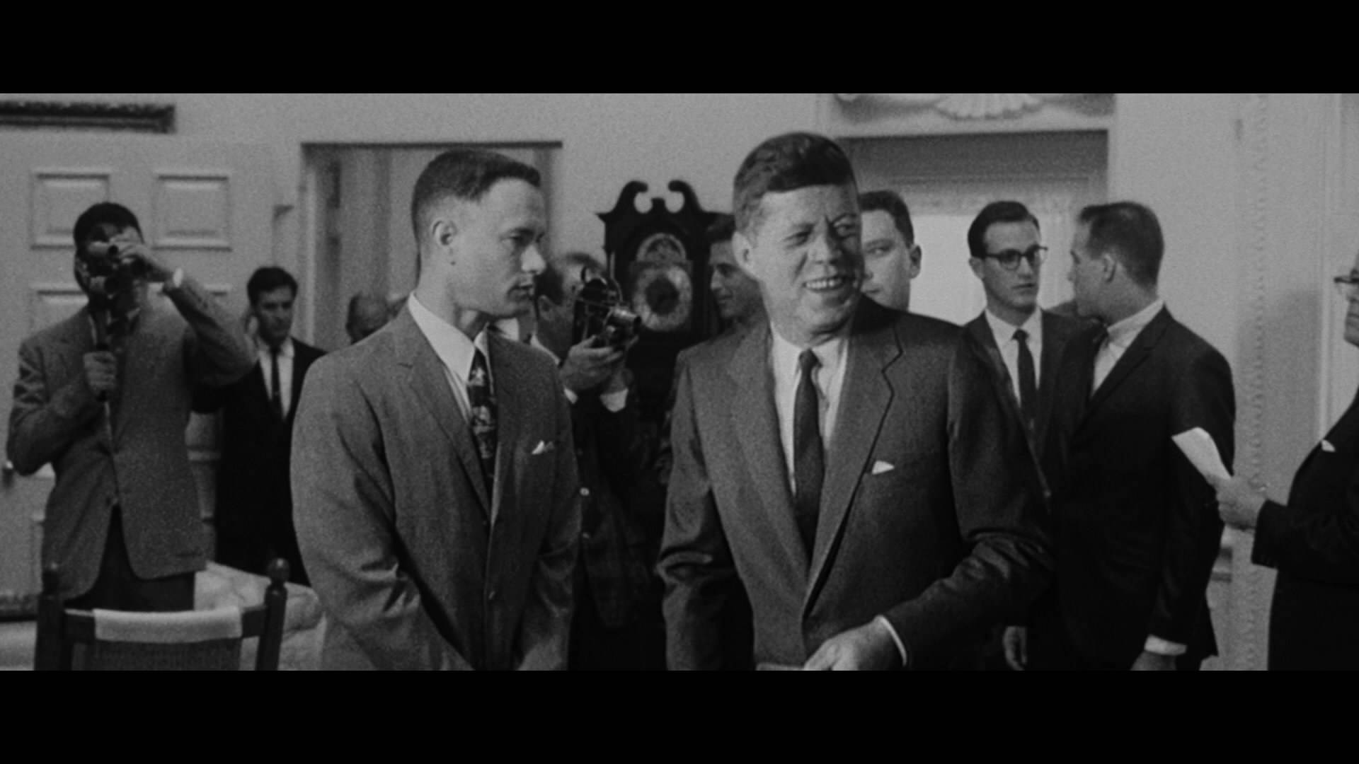 阿甘與約翰·肯尼迪 壁紙 — JFK 在正式室內環境中被記者及攝影師包圍，溫暖微笑的黑白照片。