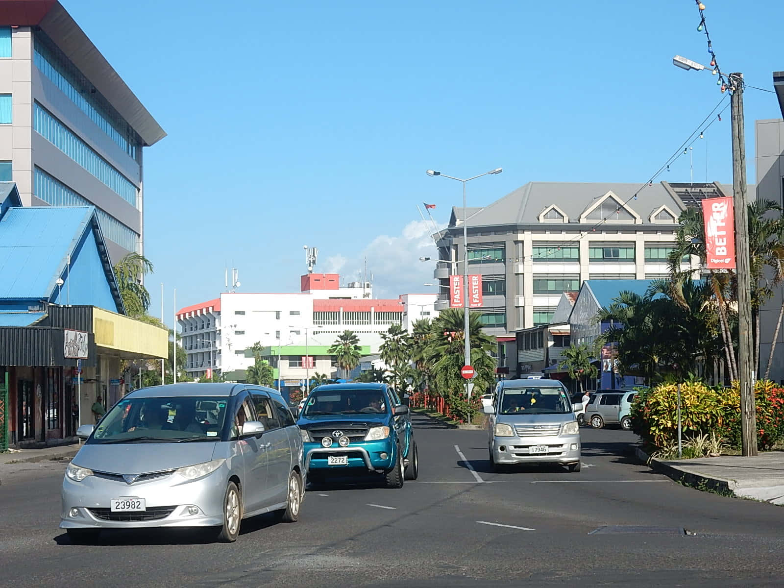 汽車喺陽光明媚嘅熱帶城市街道上行駛，兩旁種滿棕櫚樹同現代建築。