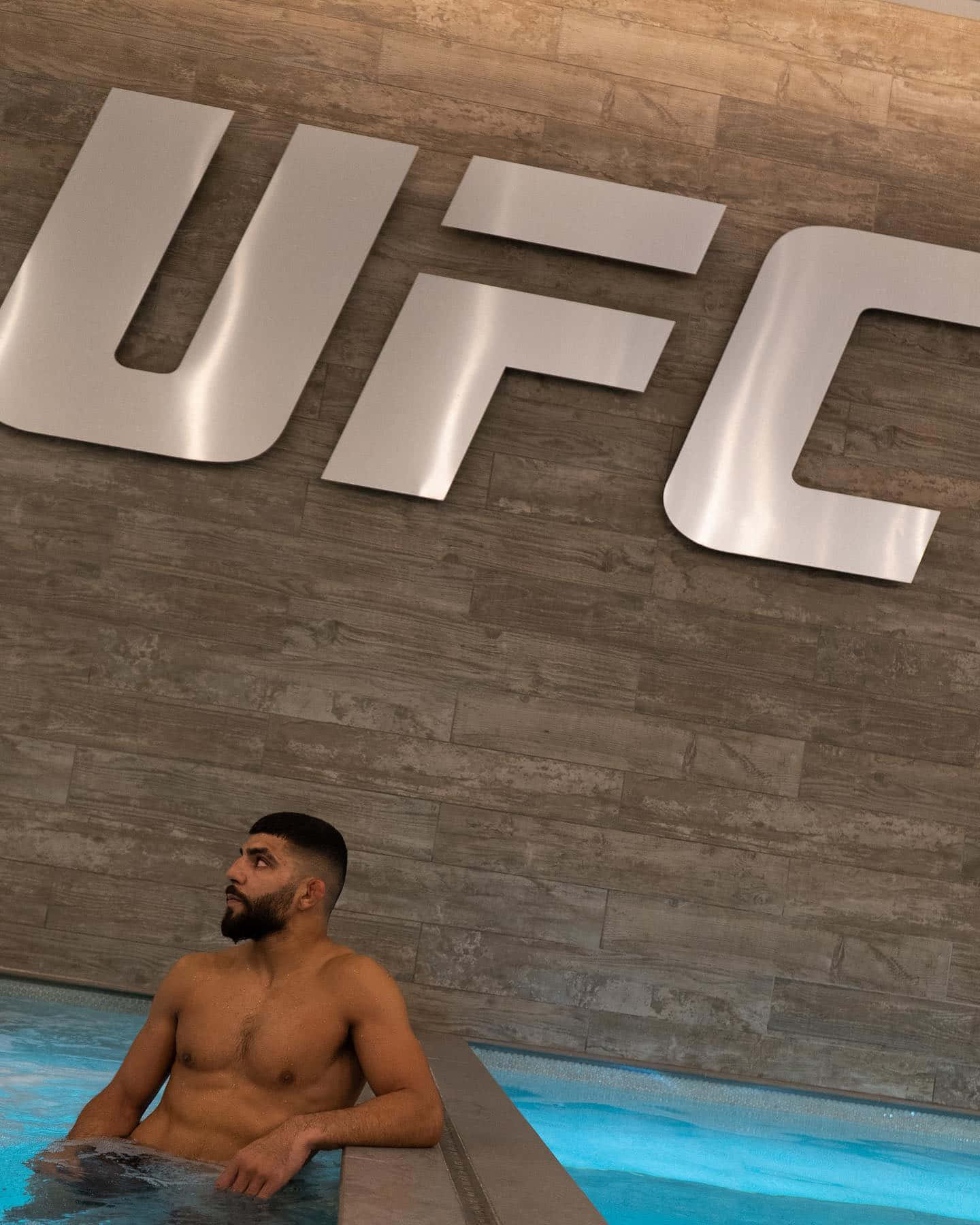 一個留鬍子的男人在木牆上一個大型UFC標誌下的藍色水池中放鬆。