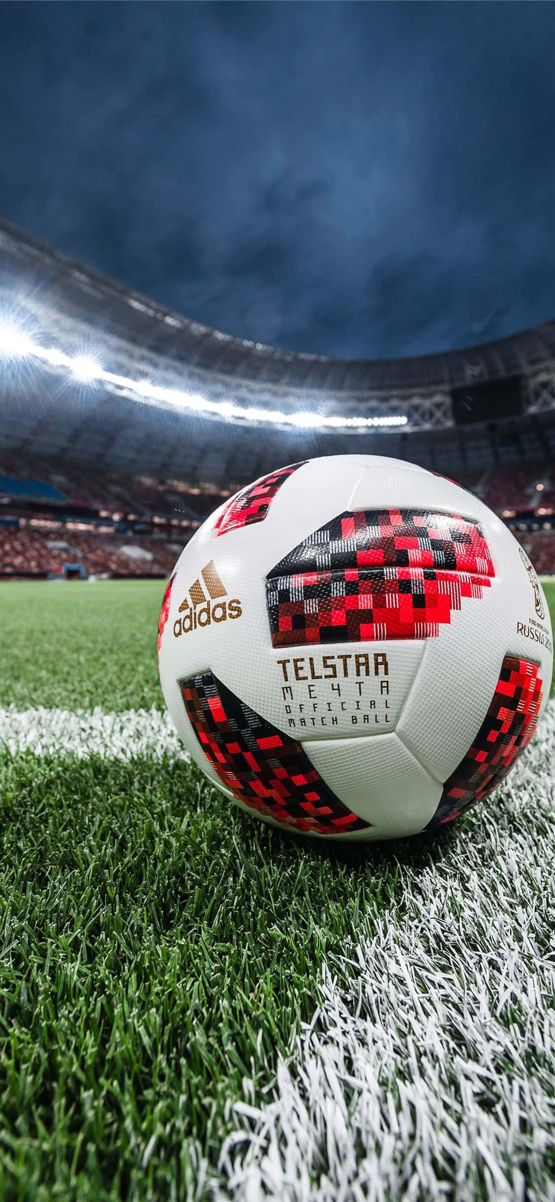 阿迪達斯Telstar足球背景 壁紙 — 一個 Telstar 18 足球放在充滿活力的綠色草地上，背景是球場燈光下模糊的人群。
