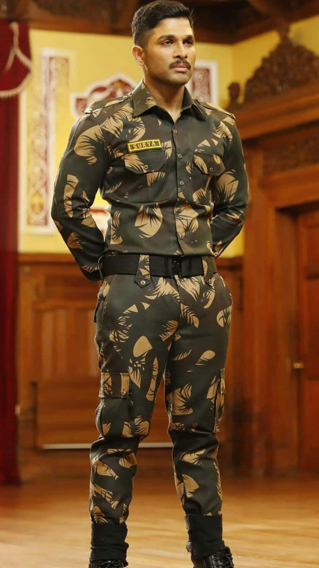 阿魯·阿俊 - 「戰士蘇里亞」全情投入動作狀態 壁紙 — 一名身穿葉紋軍服的男子在木質室內自信地雙手放在背後站立。