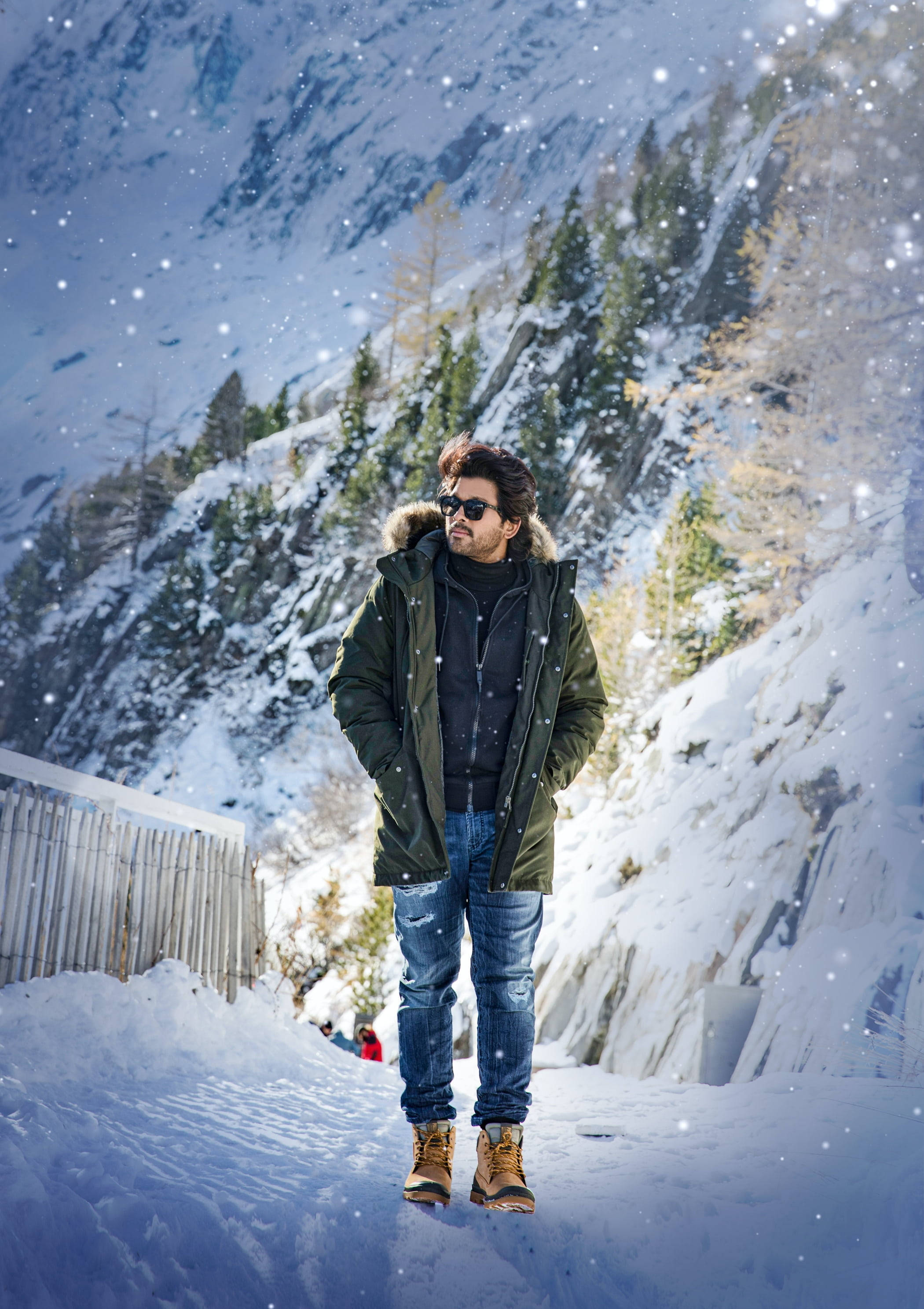 阿魯·阿爾俊高清冬季雪景 壁紙 — 一名穿著綠色冬季外套的男子自信地站在飄雪中，背景是白雪覆蓋的山脈和常青樹。