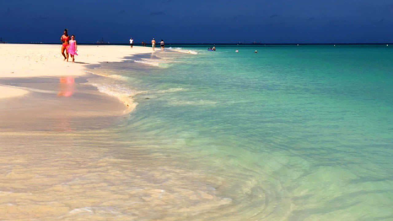阿魯巴海灘寧靜午後 壁紙 — 充滿活力的熱帶海灘場景，碧綠的海水拍打著白色沙灘，人們在深藍色的天空下沿著海岸線漫步。