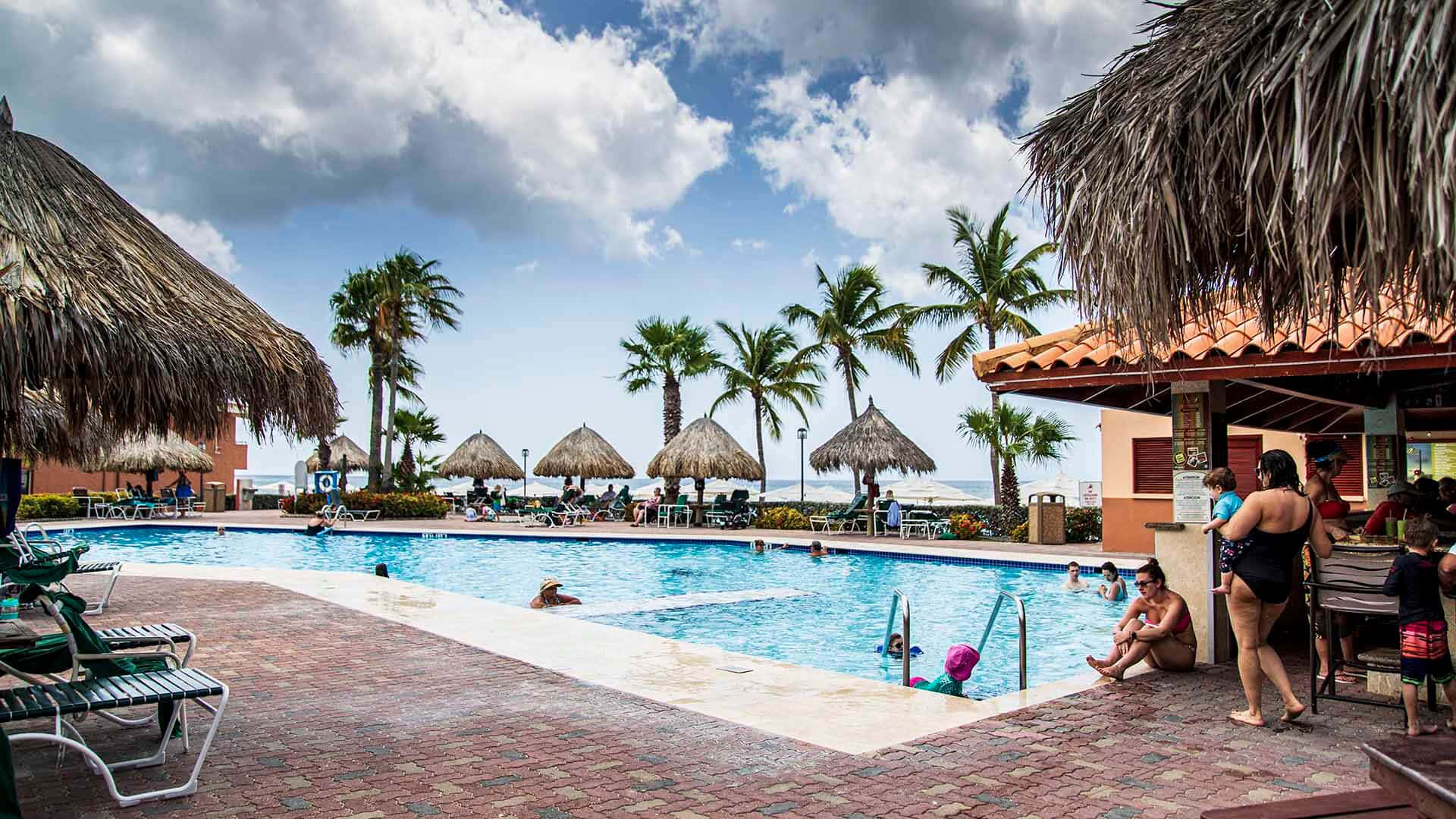 阿魯巴海灘與游泳池照片 壁紙 — 一個充滿活力的度假村泳池，周圍環繞著棕櫚樹和茅草傘，天空部分多雲。