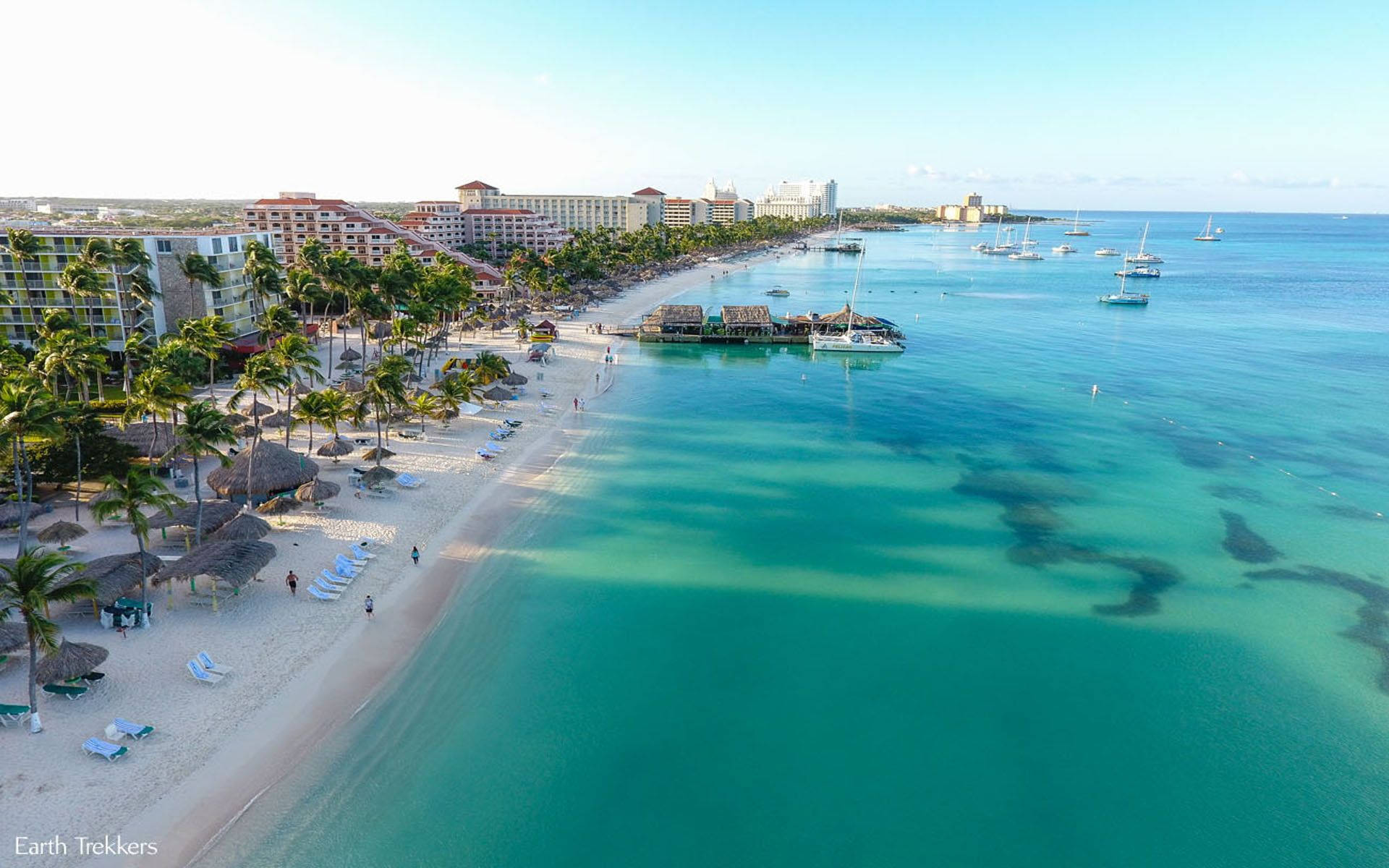 阿魯巴艾爾棕櫚海灘 壁紙 — 熱帶海灘的鳥瞰圖，有碧綠的海水、棕櫚樹、度假村建築和帆船，天空晴朗湛藍。