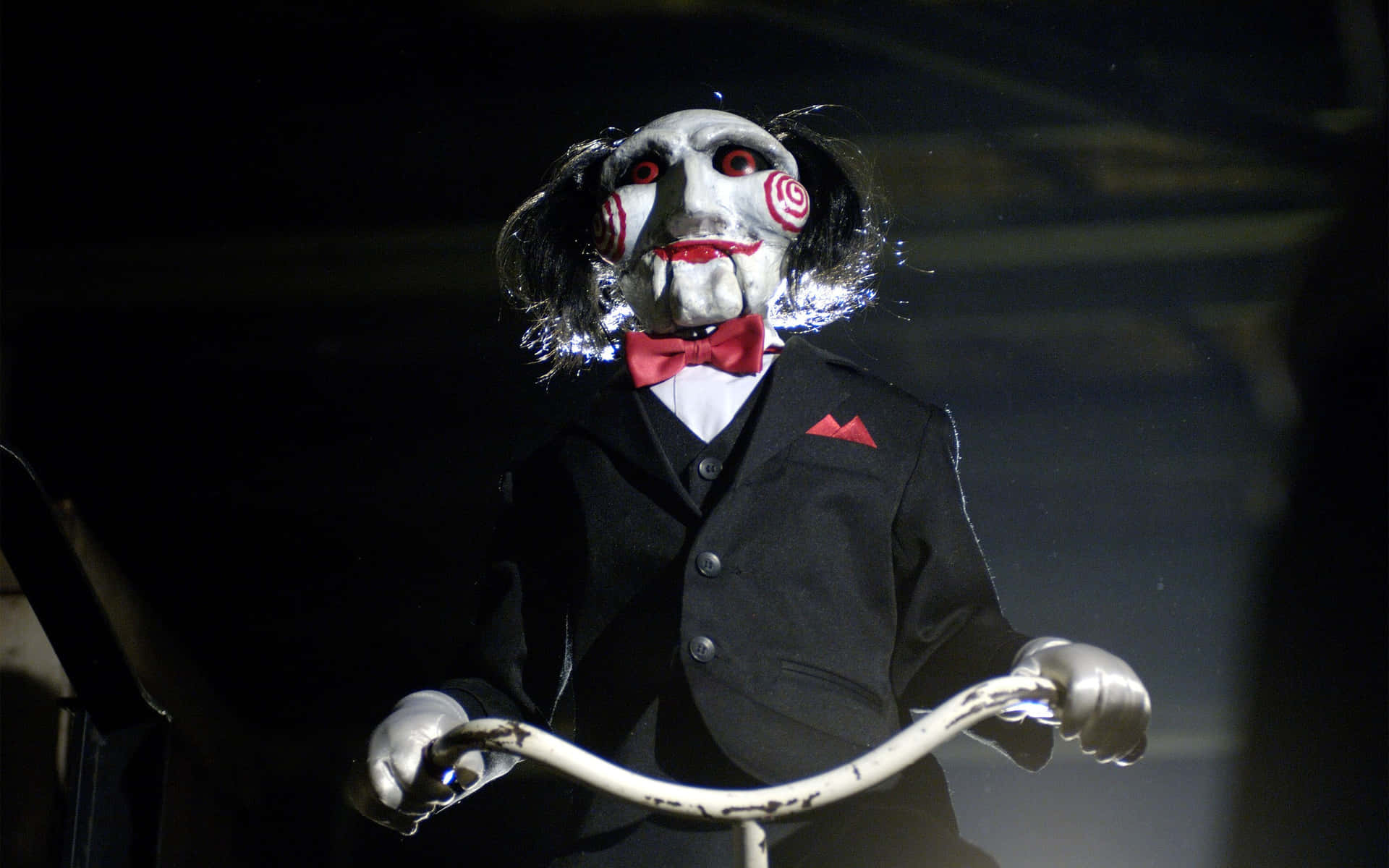 恐怖木偶穿著黑色西裝和紅色領結，在聚光燈下握著自行車把手。