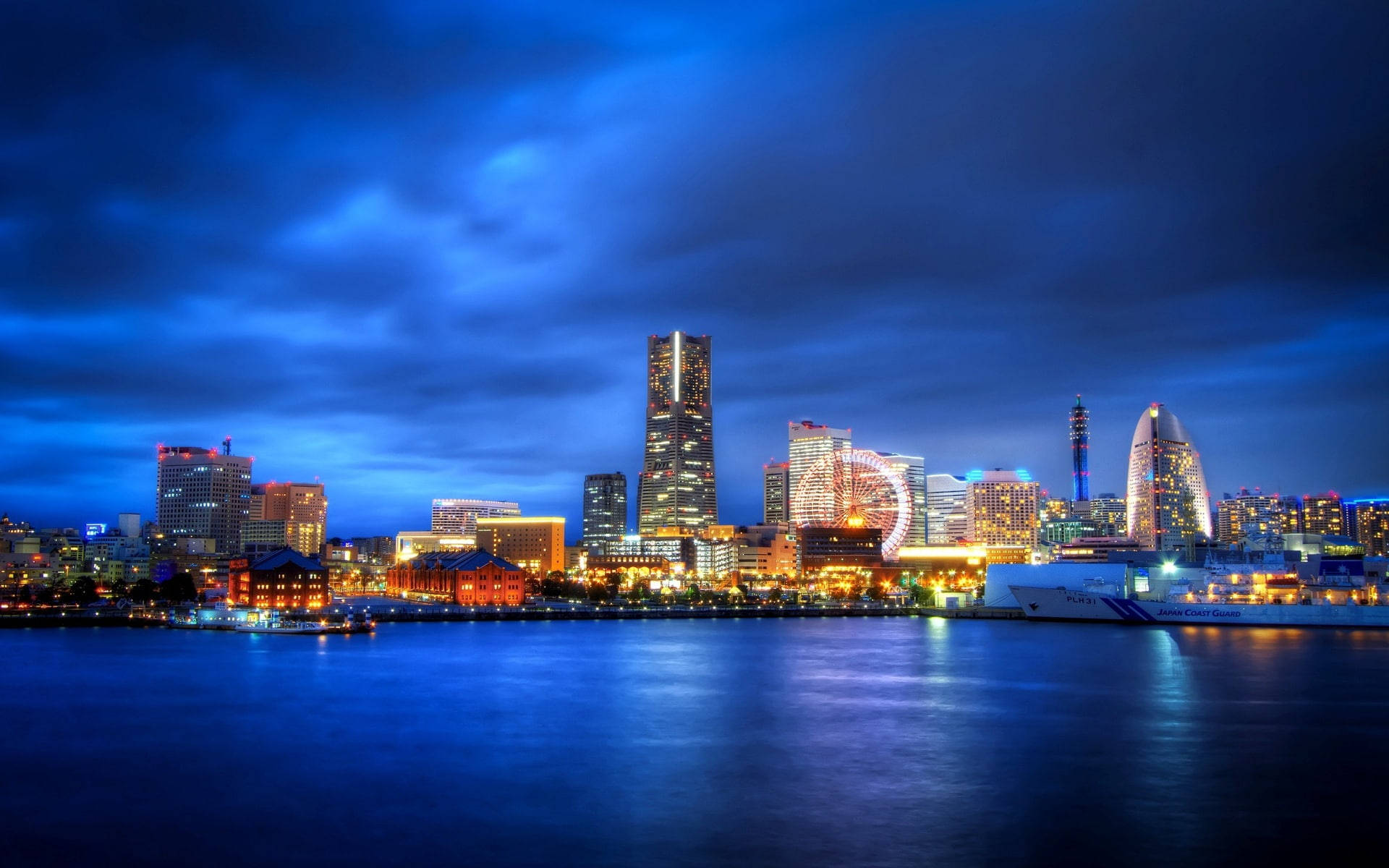 陰沉天色下的橫濱城市景觀 壁紙 — 黃昏時分充滿活力的城市天際線，閃爍的摩天大樓和摩天輪倒映在平靜的水面上，天空深藍多雲。