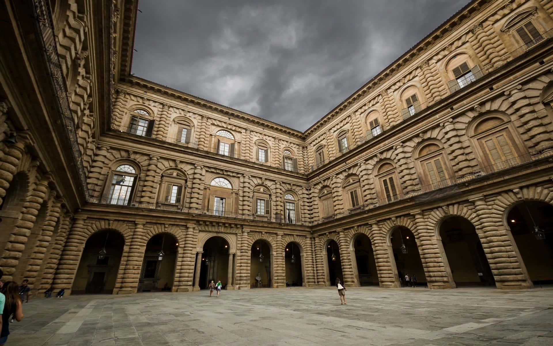 陰雲密布下的皮蒂宮 壁紙 — Palazzo Medici Riccardi 嘅宏偉石頭庭院，喺戲劇性嘅暴風雲下，有幾個訪客喺拱形柱廊下行走。