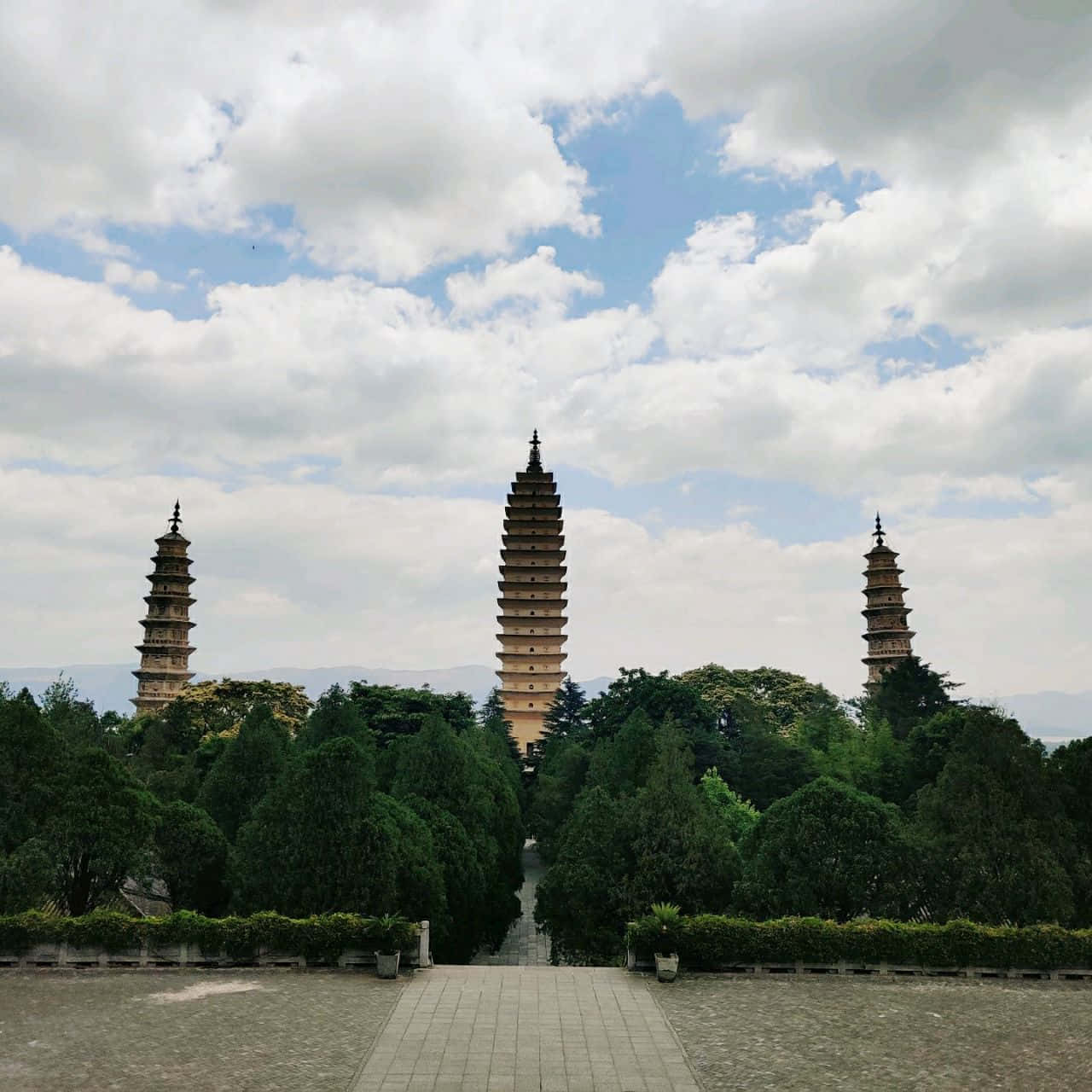 陰雲密布的三塔 壁紙 — 三座古老石塔聳立在茂密綠樹之上，天空部分多雲，一條鋪砌小徑通向它們。