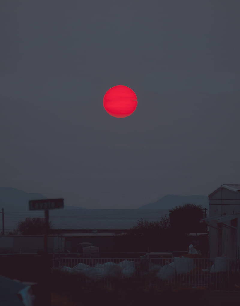 紅色的太陽在海邊小鎮落下，建築物和山丘形成剪影。