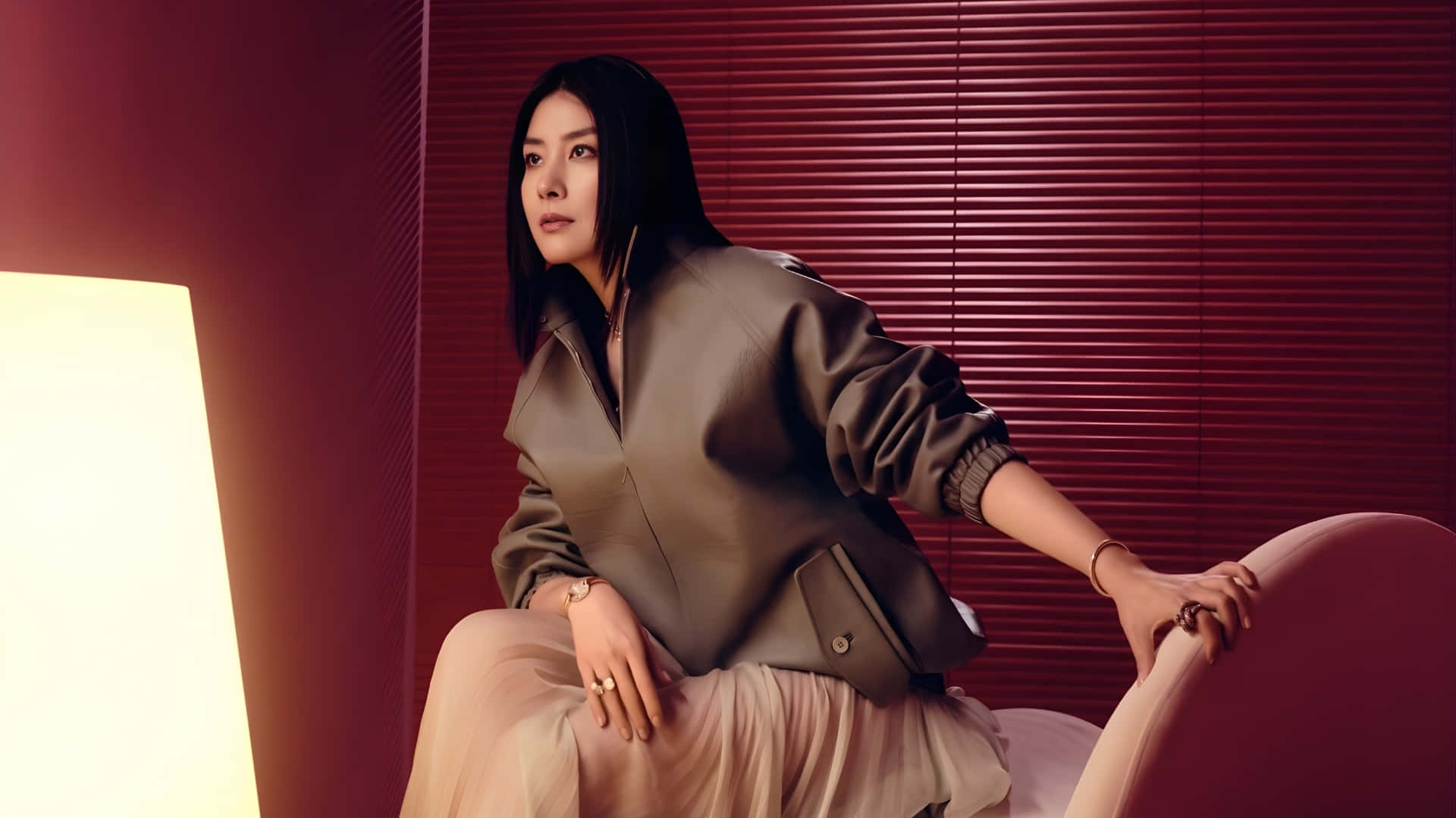 陳凱琳時尚現代肖像 壁紙 — 一位身穿時尚西裝外套的優雅女士，坐在一張弧形椅子上，背景是深紅色的條紋牆，沐浴在溫暖的環境光中。