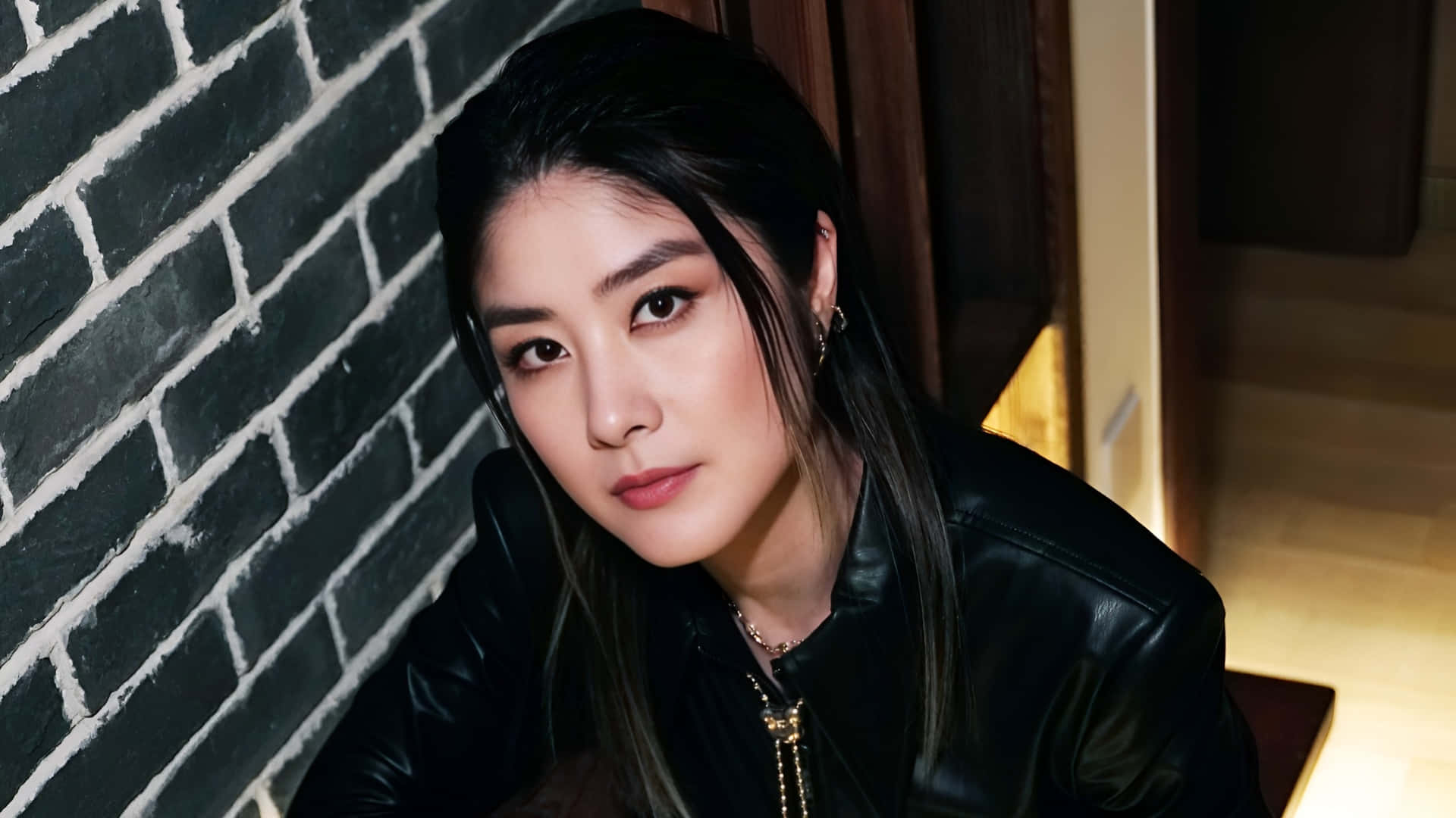 陳凱琳時尚肖像 壁紙 — 一位深色頭髮、眼神銳利的女士，在溫暖的室內燈光下，穿著黑色皮夾克靠著黑色磚牆。