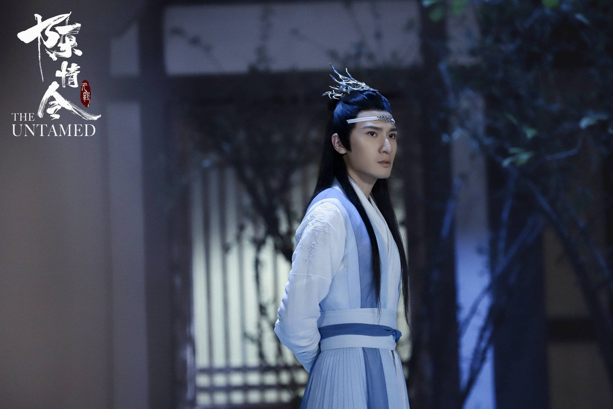 《陳情令》左惟君 壁紙 — 一名身穿傳統藍白長袍的年輕男子夜晚站在燈光昏暗的庭院中，長長的黑髮上戴著銀色頭飾。