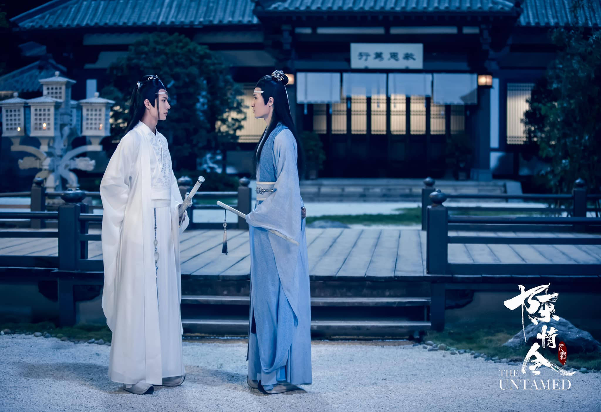 《陳情令》蘭家雙璧 壁紙 — 兩個身穿飄逸白色和藍色長袍的男人在一個寧靜的黃昏庭院中的木橋上面對面站立，周圍是傳統建築。