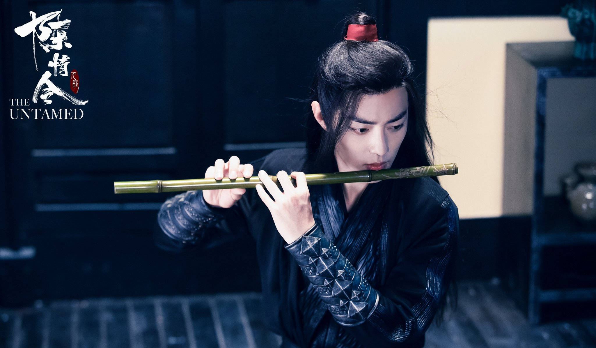 《陳情令》魔修魏無羨 壁紙 — 一位身穿深色傳統長袍的年輕人在昏暗的房間裡專注地吹奏竹笛。