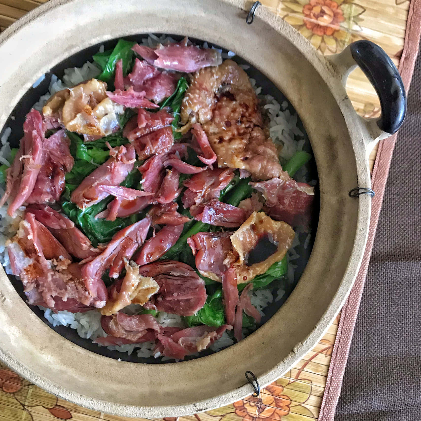 陶鍋鴨肉飯配滷鴨腿 壁紙 — 一個冒著蒸氣的砂鍋，裡面裝滿了米飯，上面鋪著切片臘肉和綠色蔬菜，放在一個花卉圖案的餐墊上。