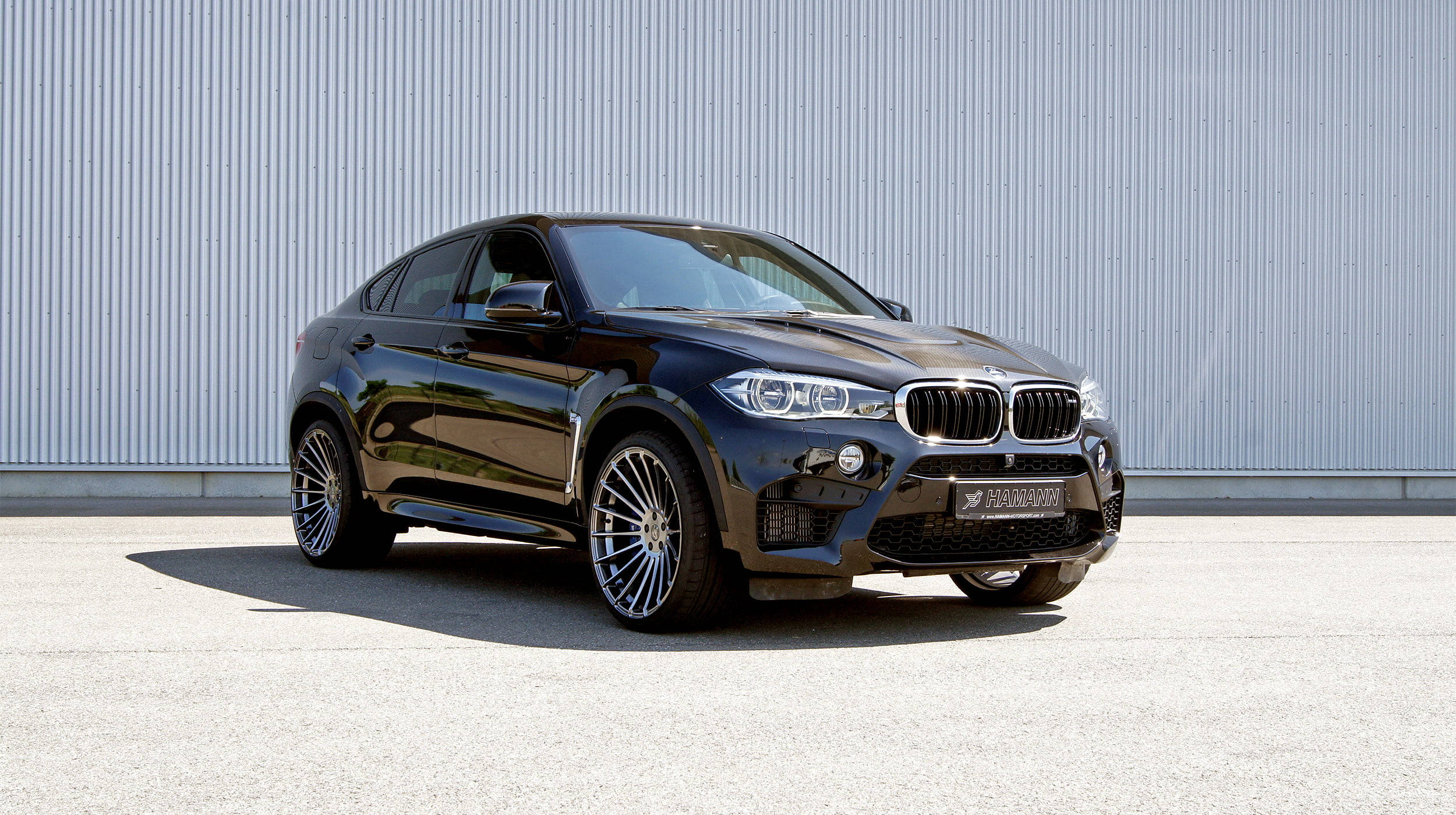 陽光下的BMW X6 M：動感與豪華的結合 壁紙 — 一輛時尚的黑色BMW X6M運動轎跑車停在混凝土上，背景是波紋金屬牆，在明亮的日光下。