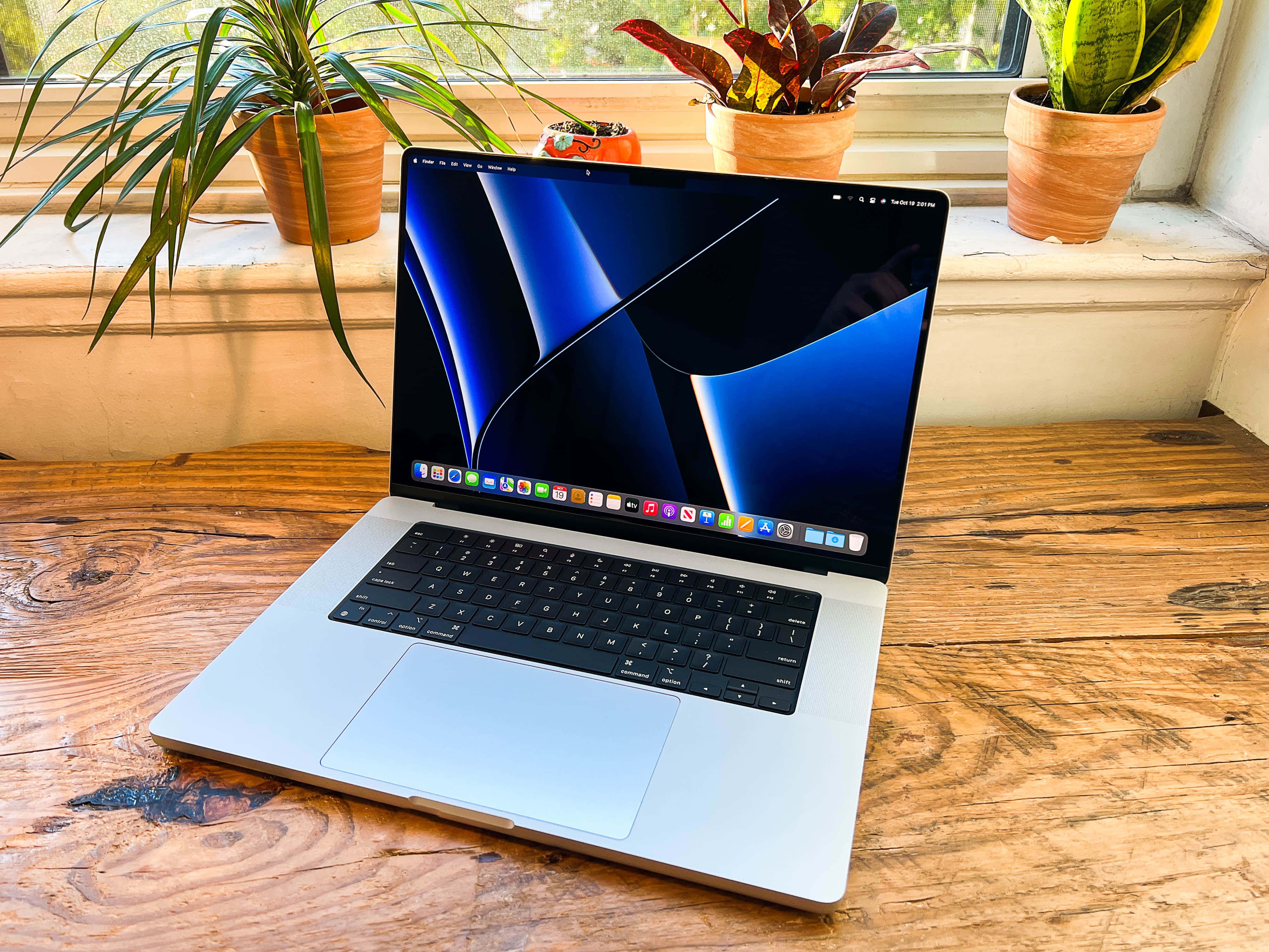 陽光下的現代 MacBook 筆記型電腦 壁紙 — 一部銀色手提電腦，帶有藍色抽象桌布，打開放在質樸的木桌上，旁邊是陽光充足的窗台，放有盆栽植物。