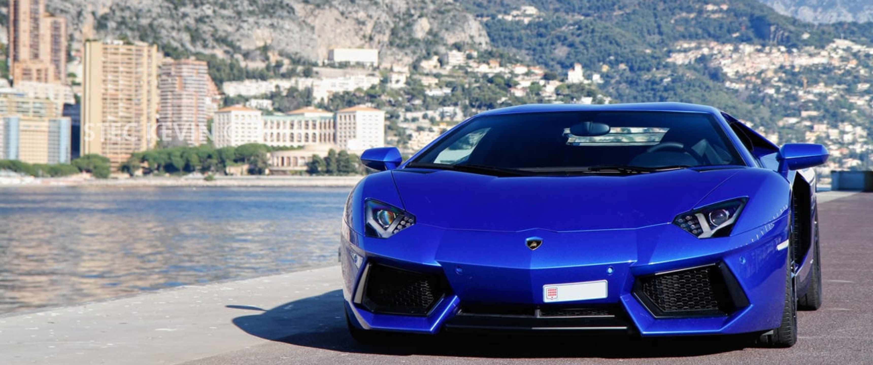 陽光下閃耀的紅色蘭博基尼跑車 壁紙 — 一架鮮藍色林寶堅尼Aventador停泊在海濱長廊上，背景是摩納哥的天際線和山脈。