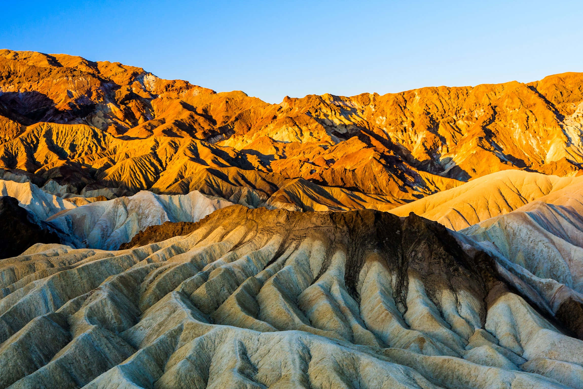 Landscapes 桌布 壁紙 — 陽光以溫暖的金色和柔和的藍色陰影沐浴著層疊的沙漠山脈，營造出戲劇性的自然景觀。